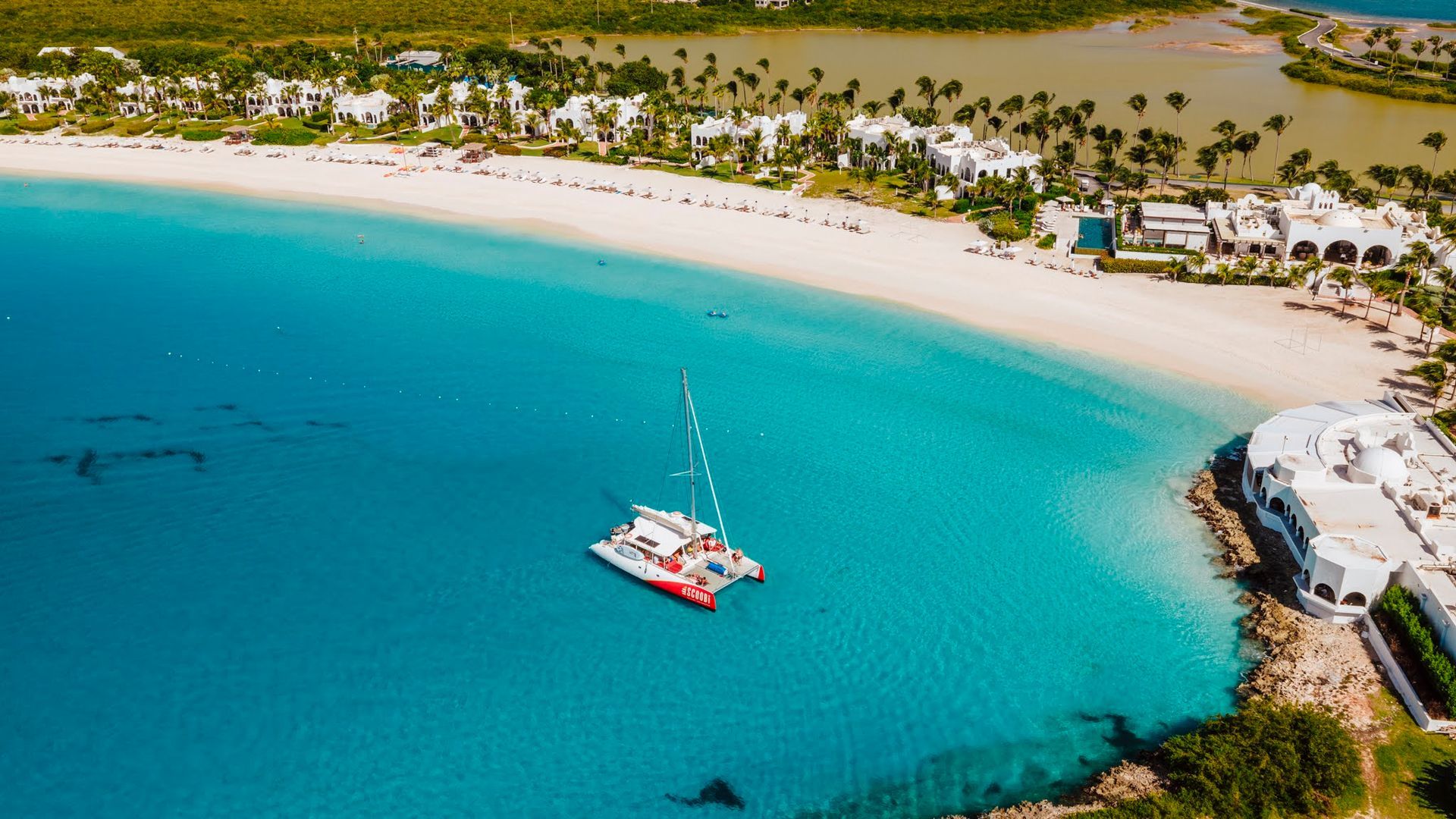 Aerial view of a white sand beach, turquoise water, and a red and white catamaran.