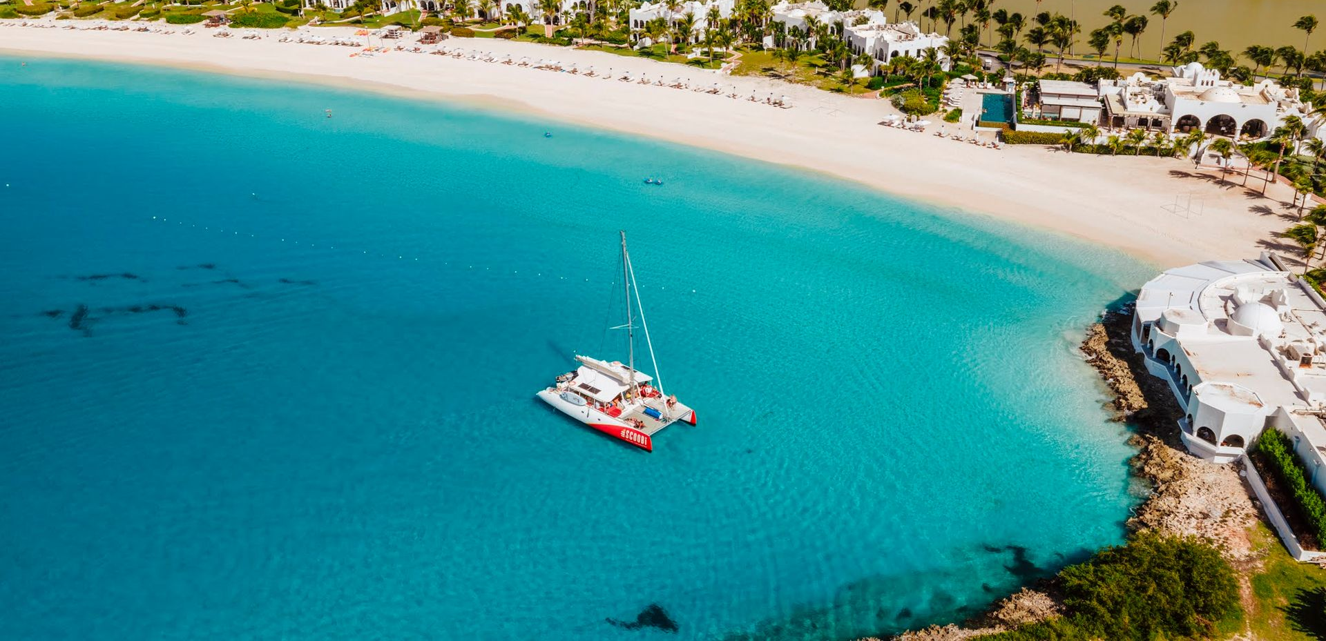 Navigation en catamaran dans des eaux turquoise près d'une plage de sable blanc bordée de complexes hôteliers.