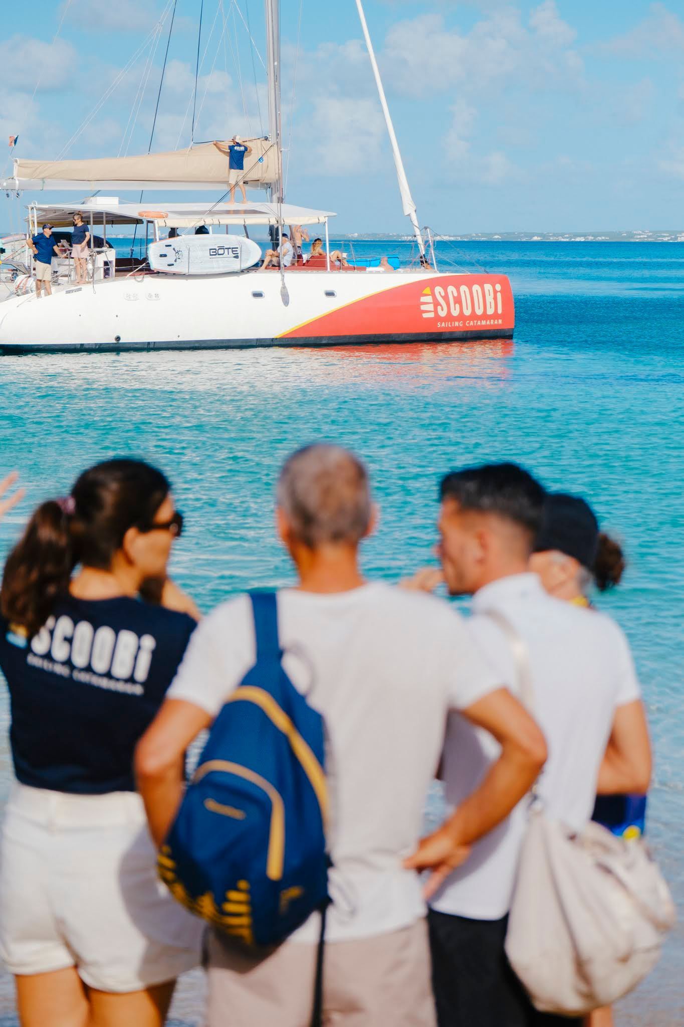 People on a beach watching a catamaran. The boat is white and orange. Clear blue water and sky.