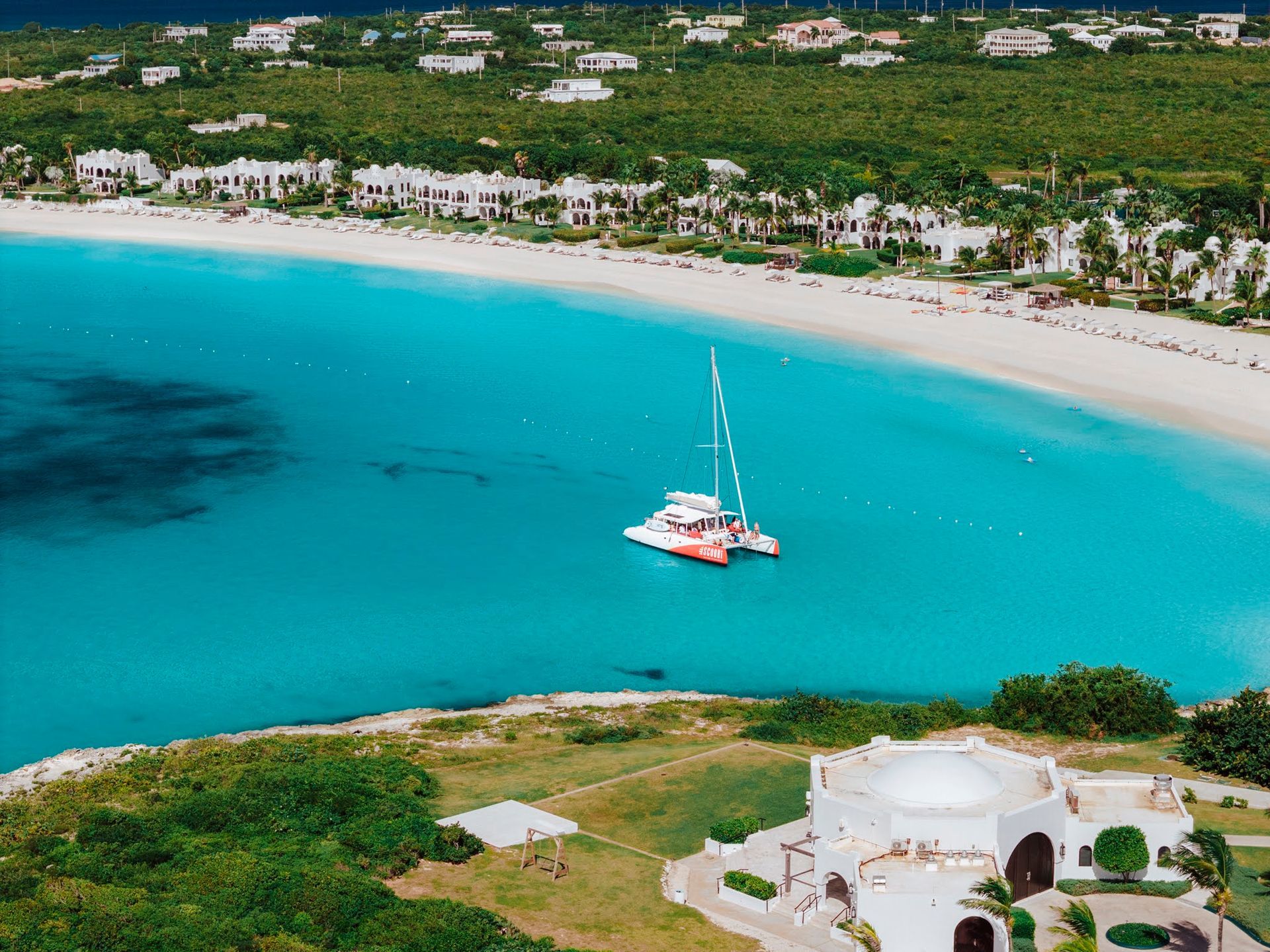 Aerial view of a turquoise bay with a white sandy beach, a catamaran, and buildings along the shore.