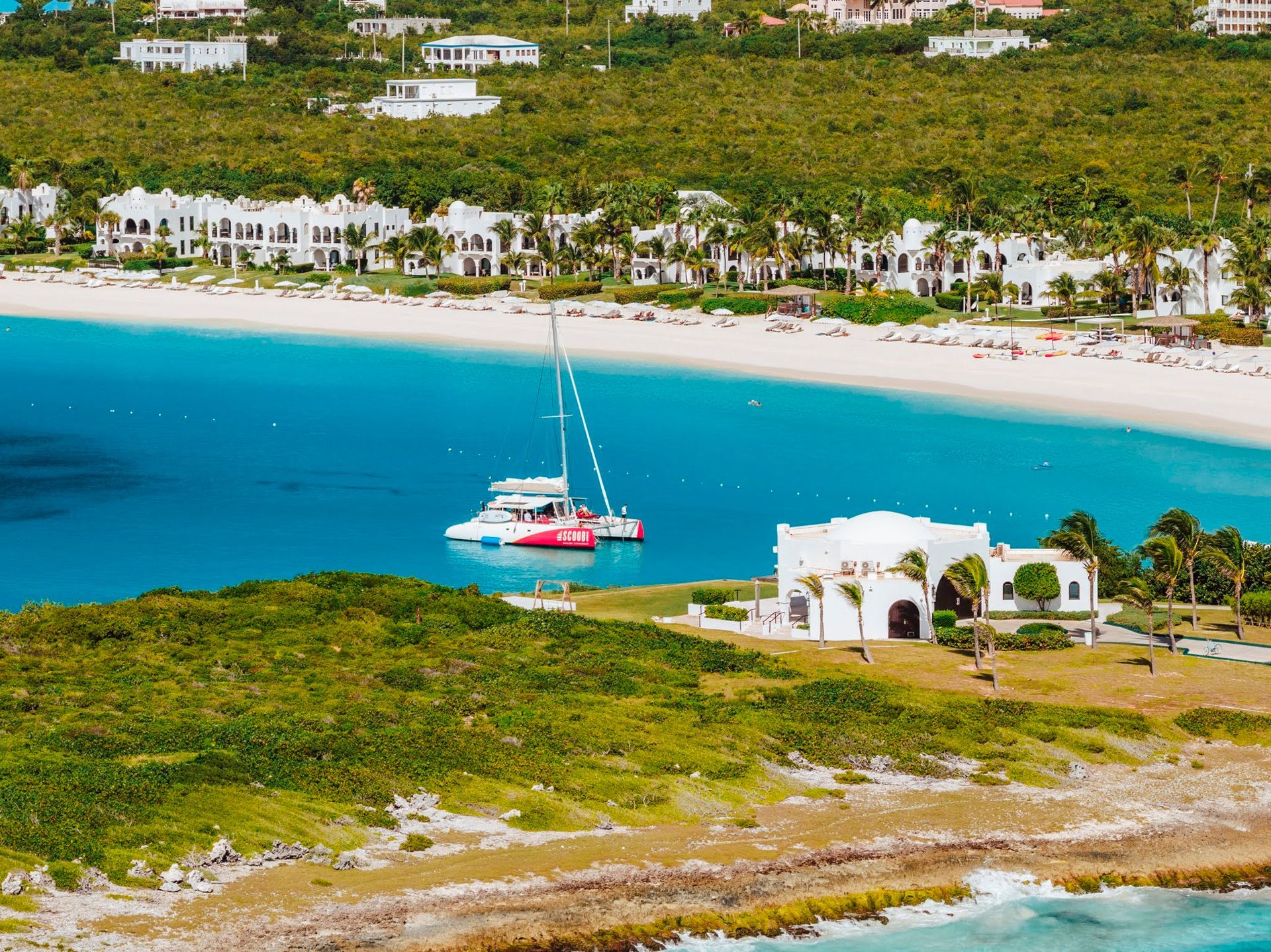 Scenic beach with turquoise water, a sailboat, white buildings, and lush green hillside.