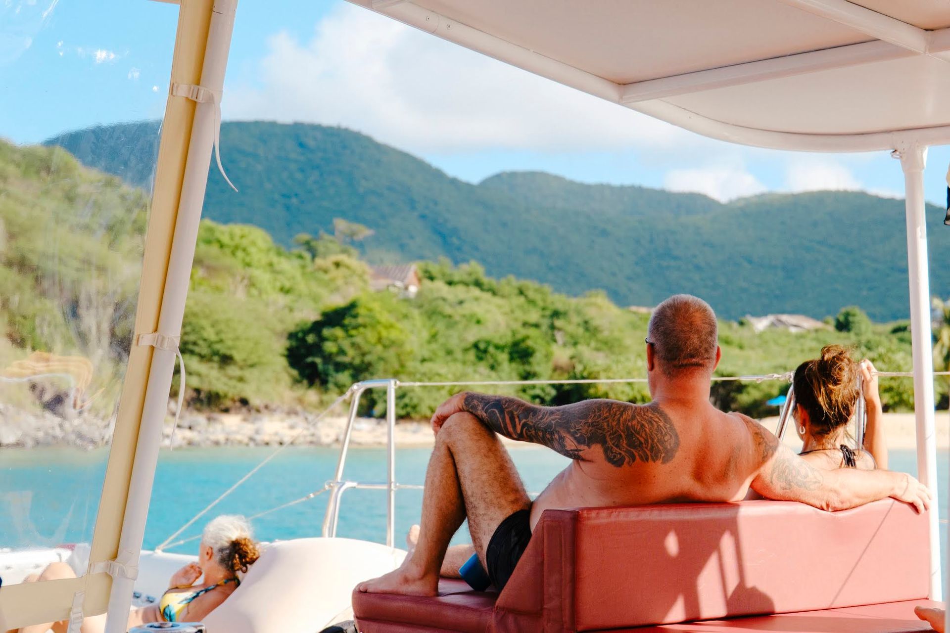 Un homme tatoué et une femme se détendent sur un bateau, avec vue sur la montagne et l'océan.