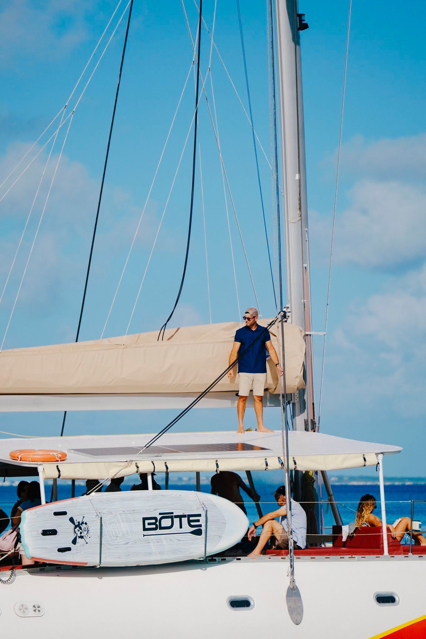 Man standing on a sailboat deck, blue sky, people relaxing nearby.