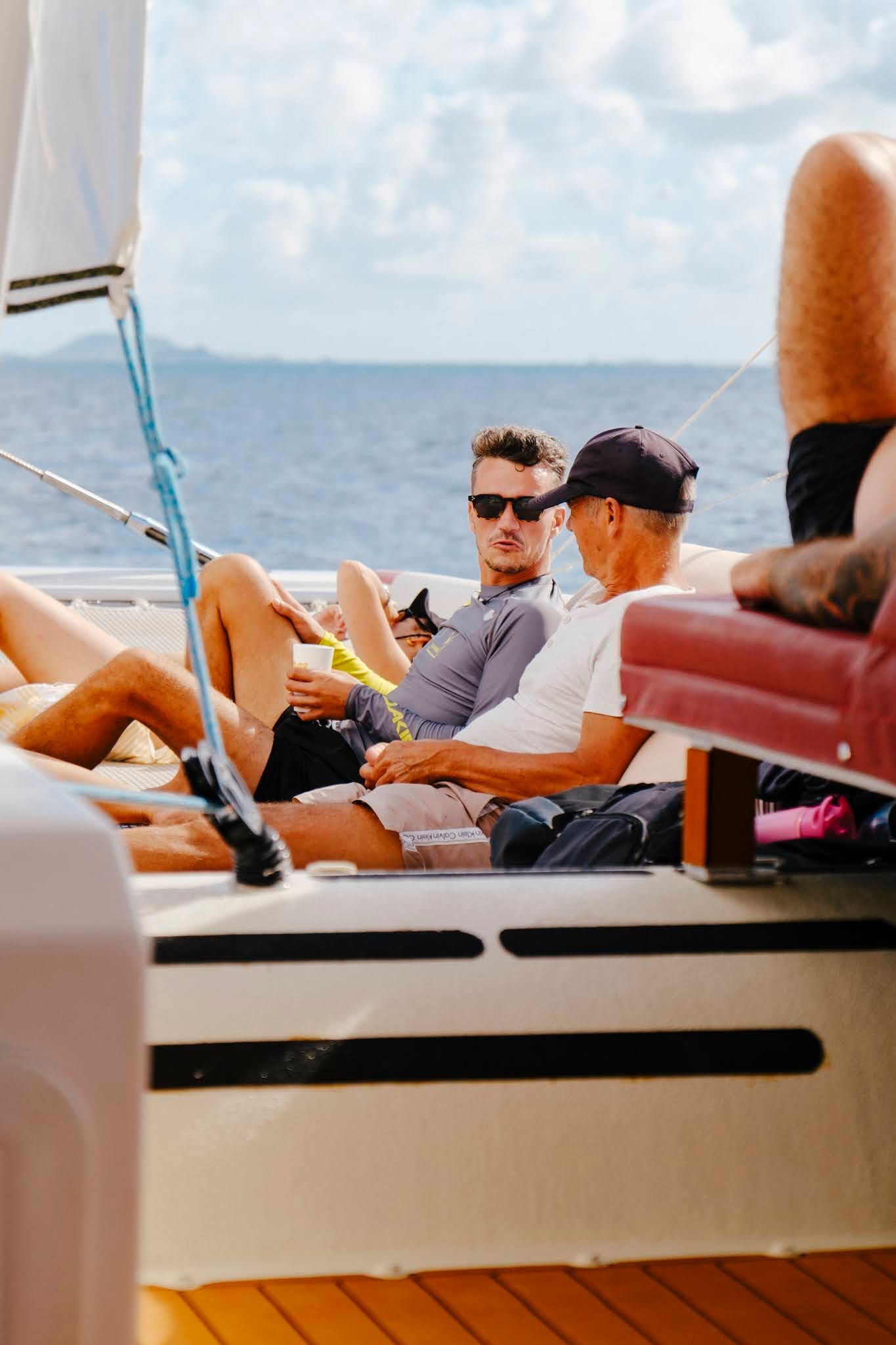 People relaxing on a sailboat, ocean in background. One wears sunglasses, another a hat. Bright sunlight.