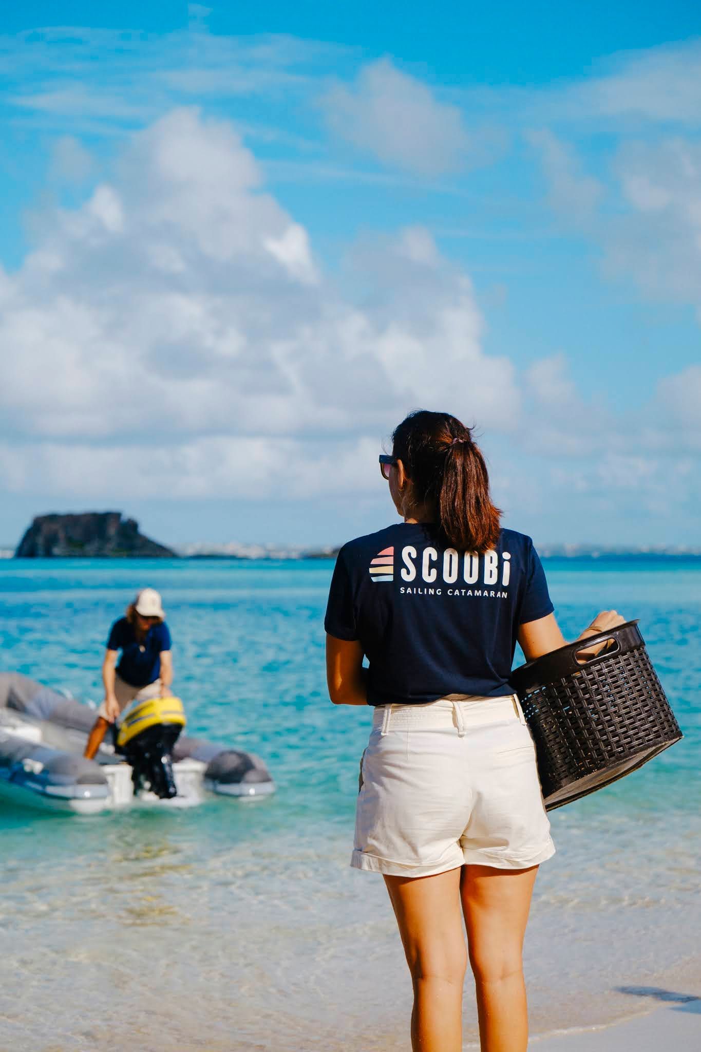 Femme sur la plage, tenant un panier, regardant vers un canot pneumatique avec une personne à bord, eau bleue, ciel ensoleillé.