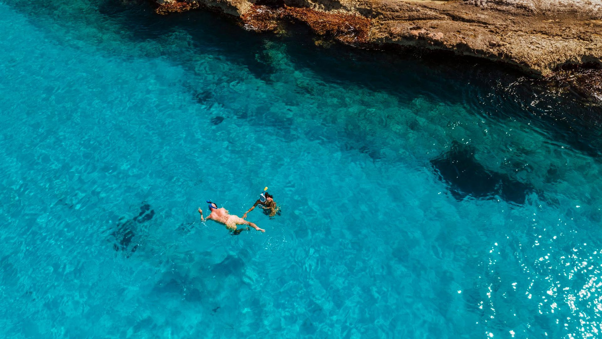 Two people snorkeling in clear turquoise water near a rocky coastline.