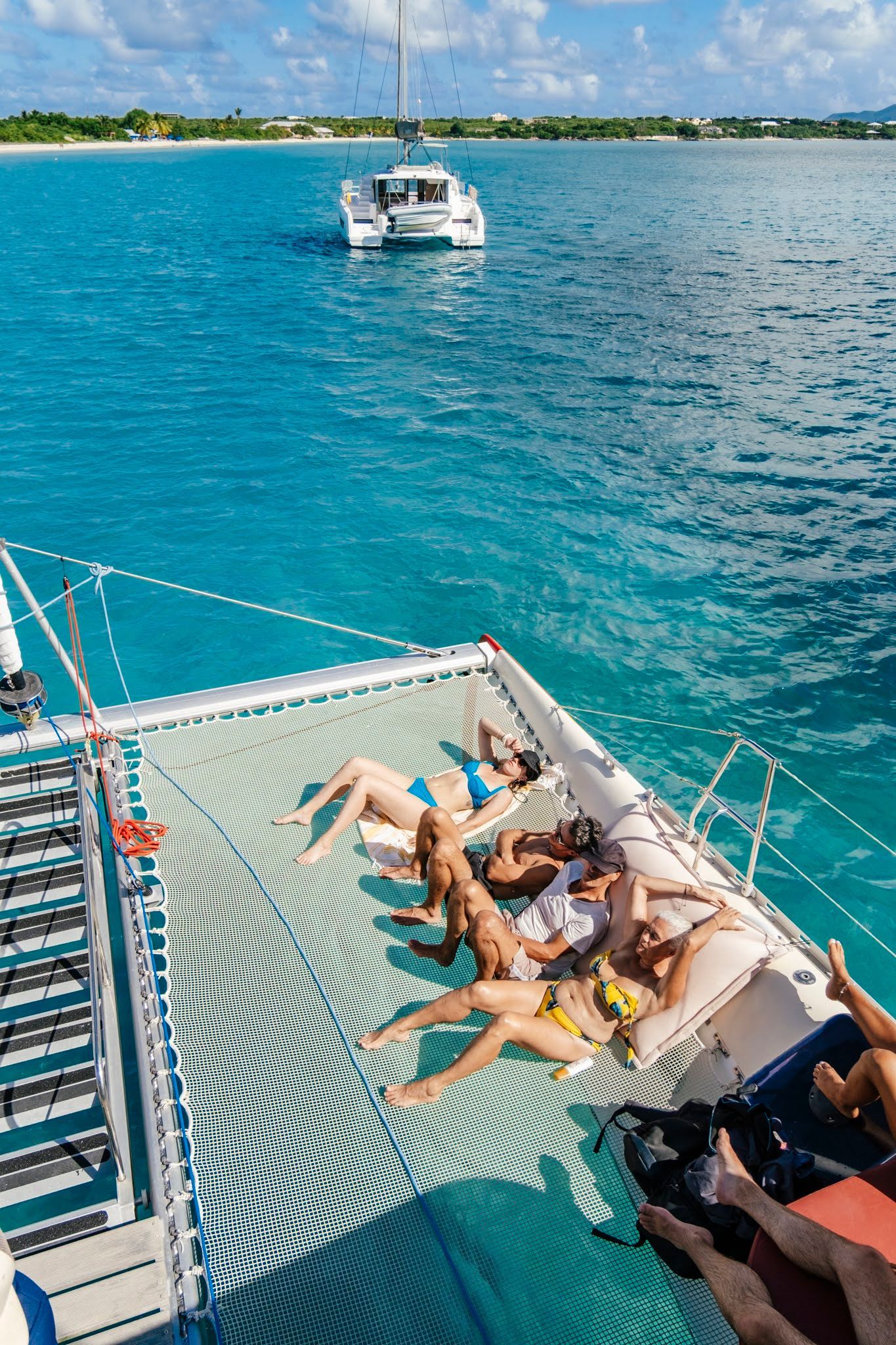 Des gens prennent un bain de soleil sur le filet d'un catamaran, au-dessus d'une eau turquoise ; un autre bateau navigue au loin.
