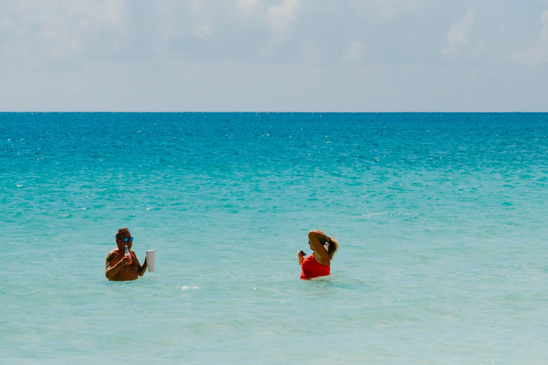 Deux personnes pataugent dans une eau turquoise ; l’une tient un objet blanc, l’autre un objet rouge. Ciel bleu au-dessus.