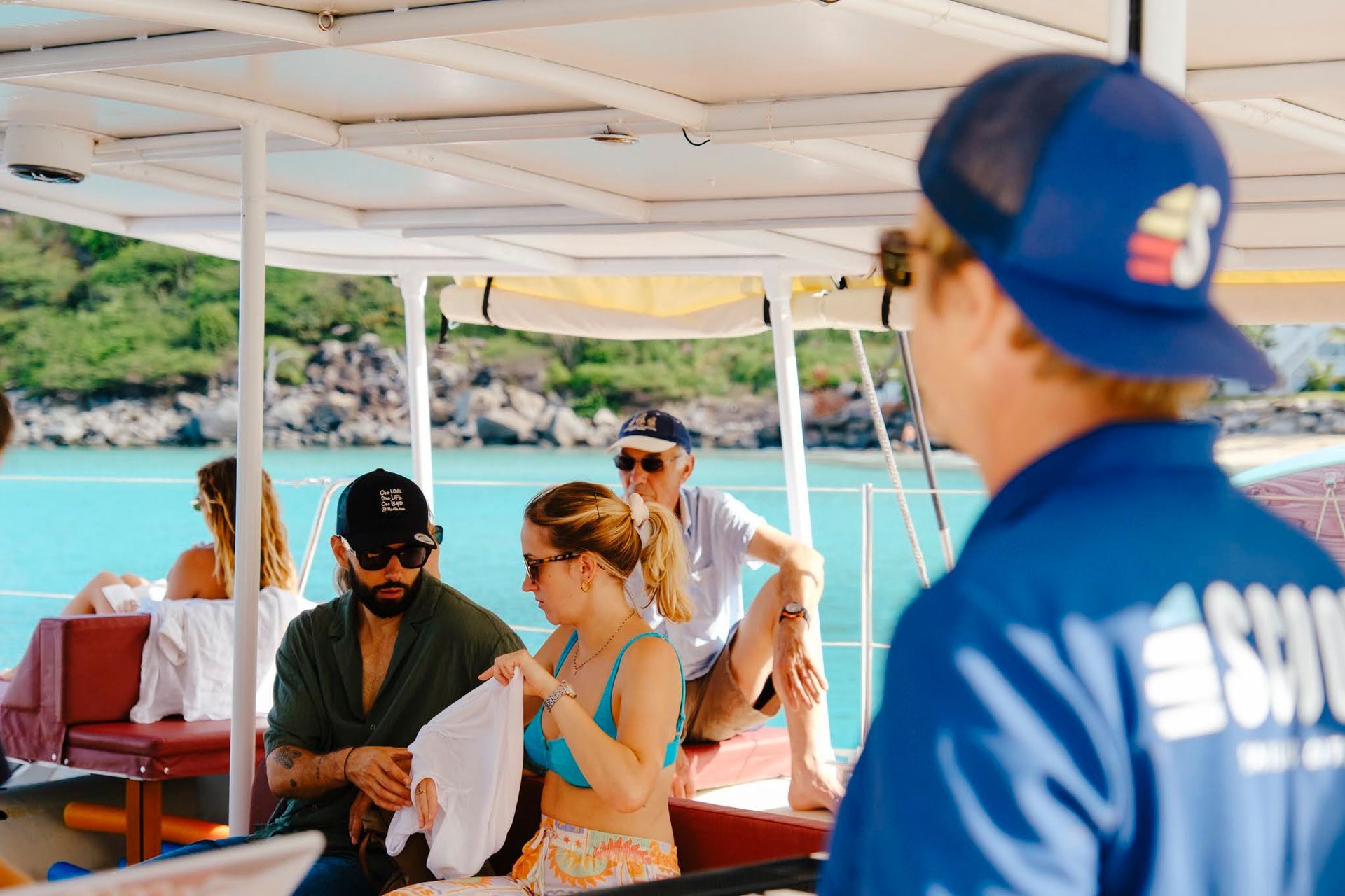 Des personnes sur un bateau près d'une plage. Une personne en uniforme bleu et chapeau ; les autres en maillot de bain et lunettes de soleil.