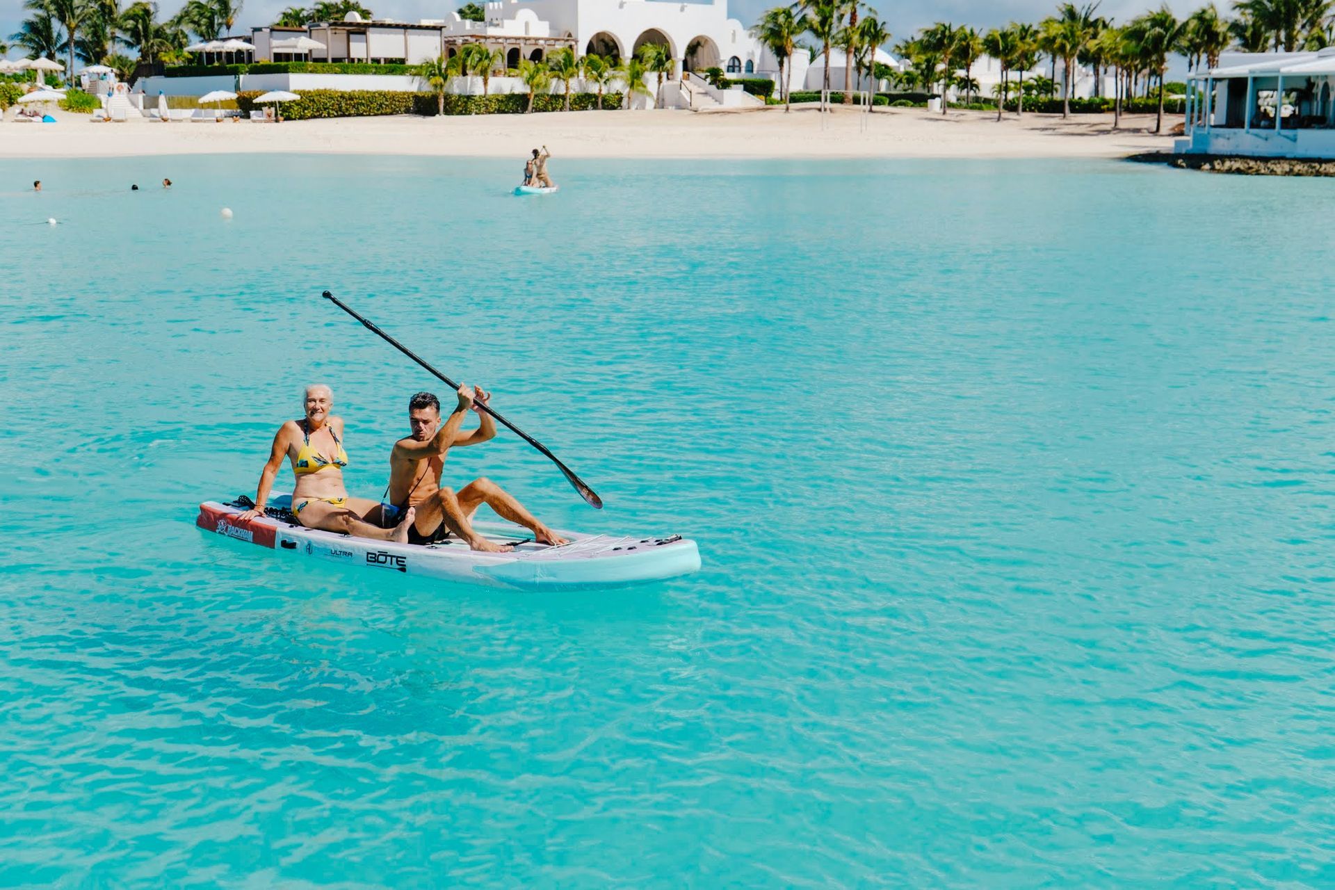 Two people on a paddleboard in turquoise water. Man paddling, woman sitting. White beach and buildings in background.