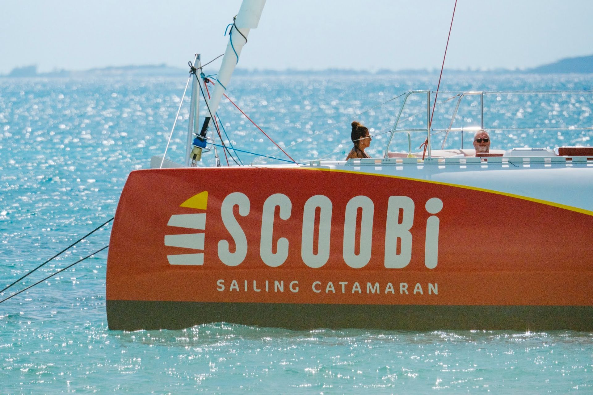 Catamaran Scoobi à voile dans une eau turquoise. Coque orange, personne sur le pont.
