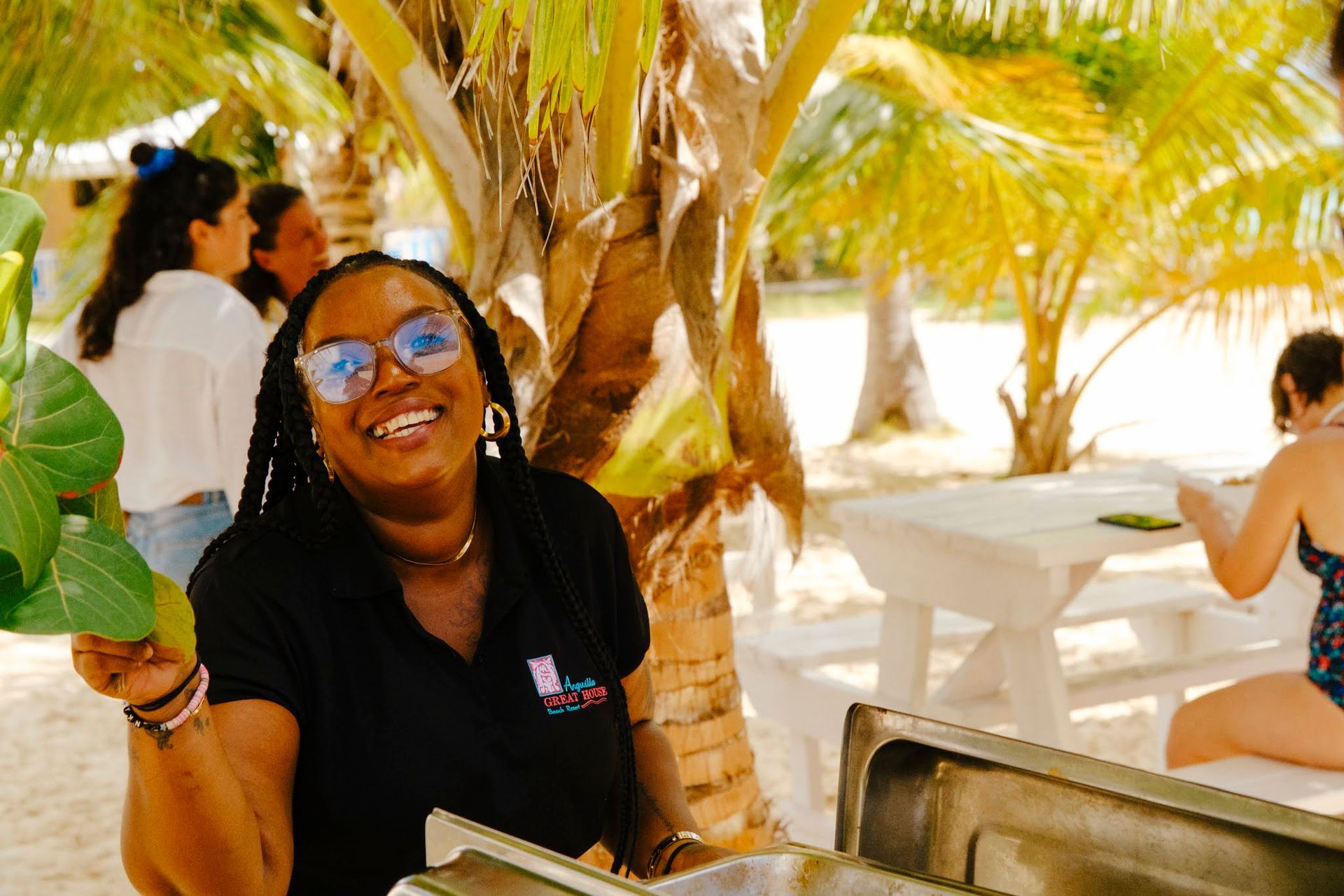 Femme souriante, tenant des feuilles, debout près d'un buffet sur une plage, sous des palmiers.