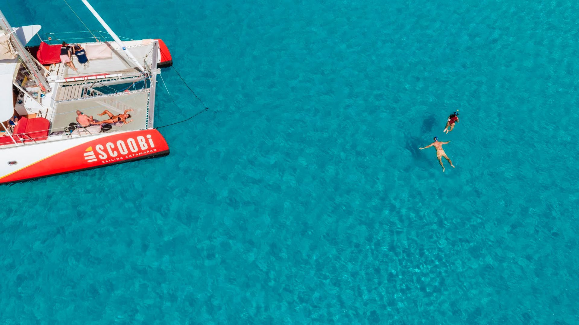 Catamaran with passengers in turquoise water, two people swimming.