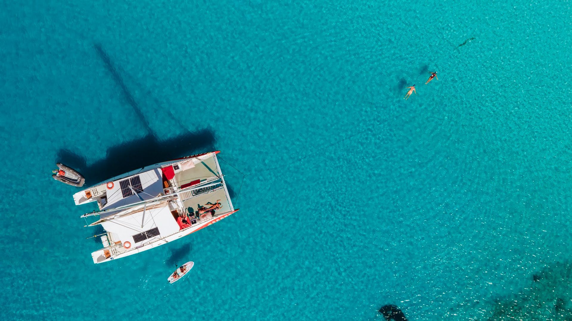 Catamaran blanc sur une eau turquoise avec des personnes à bord ; petits bateaux à proximité.