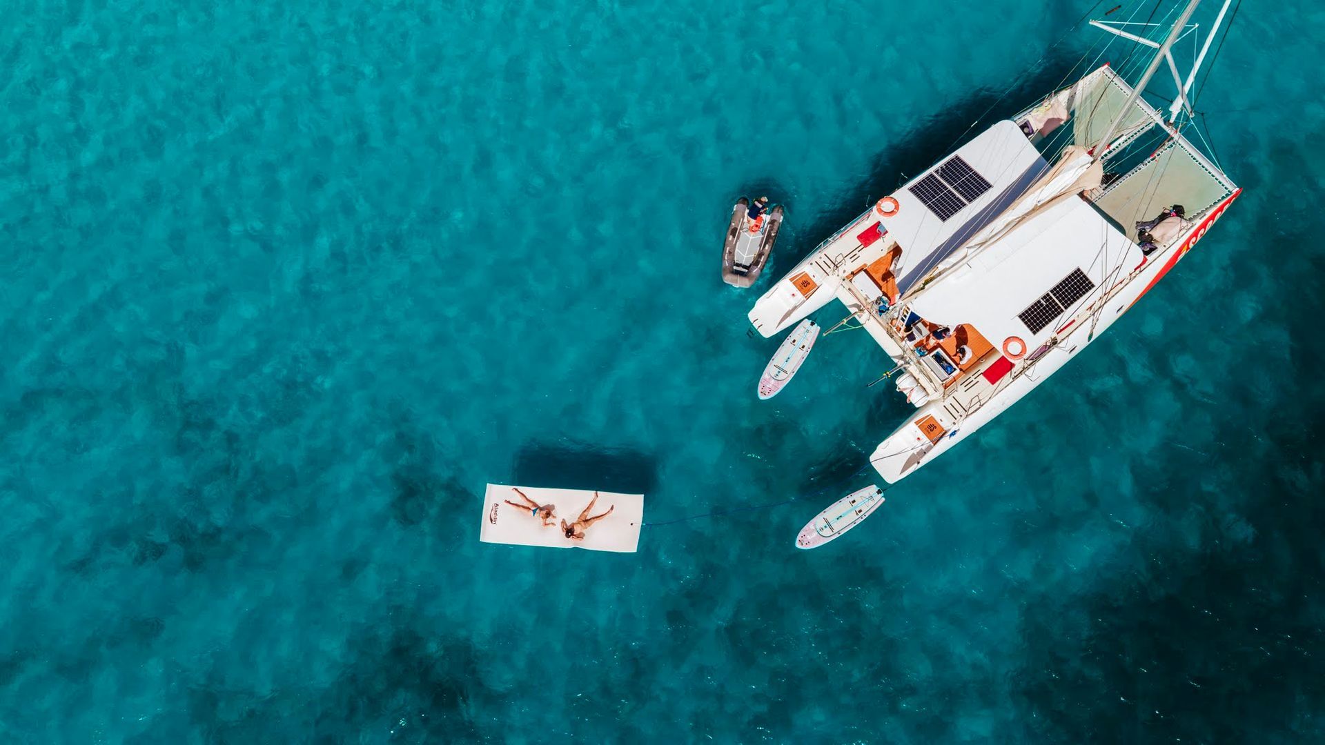 Catamaran et petite embarcation sur une eau turquoise, des personnes à bord et un radeau flottant.