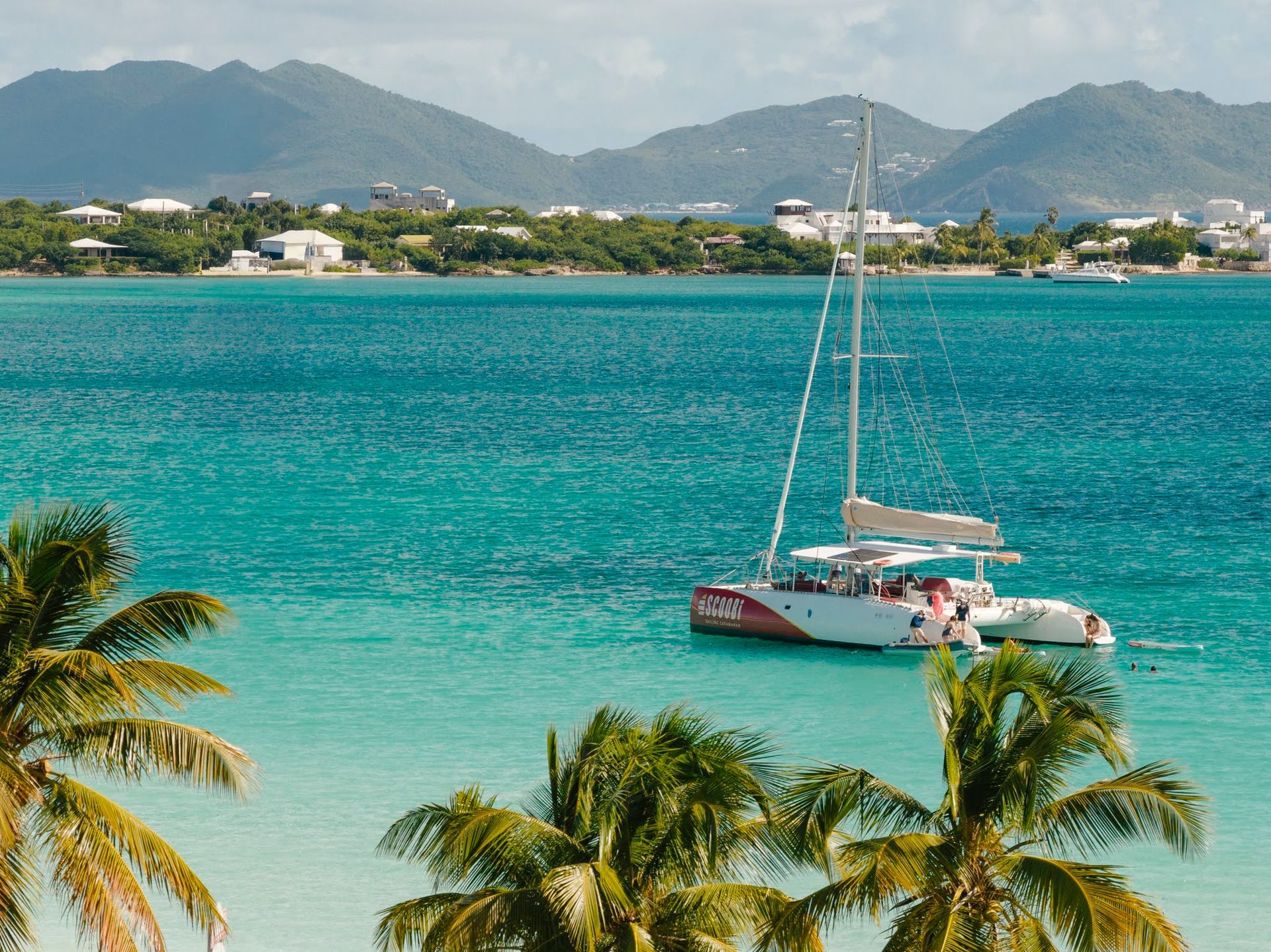 Catamaran sailing on turquoise water near a tropical island with palm trees and mountains.