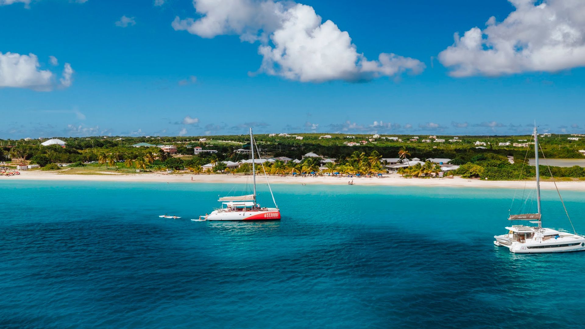Two sailboats on turquoise water near a white sand beach under a blue sky with fluffy clouds.