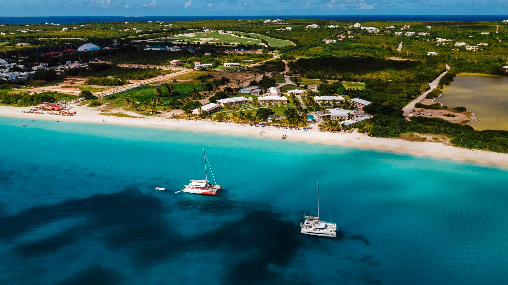 Aerial view of turquoise water and white sand beach with sailboats and buildings.