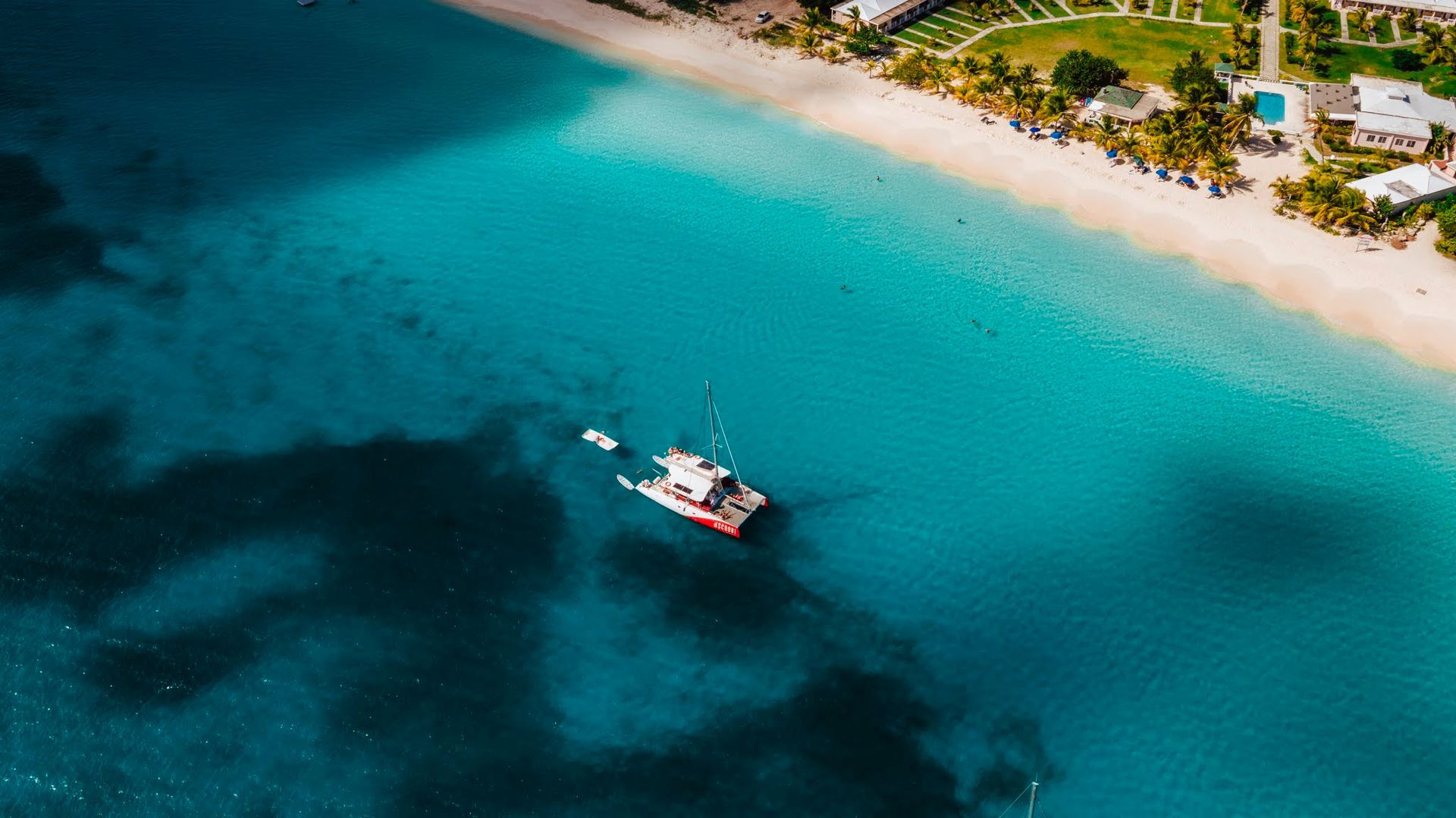 Aerial view: white sailboat in turquoise water near a white sand beach and resort with green foliage.