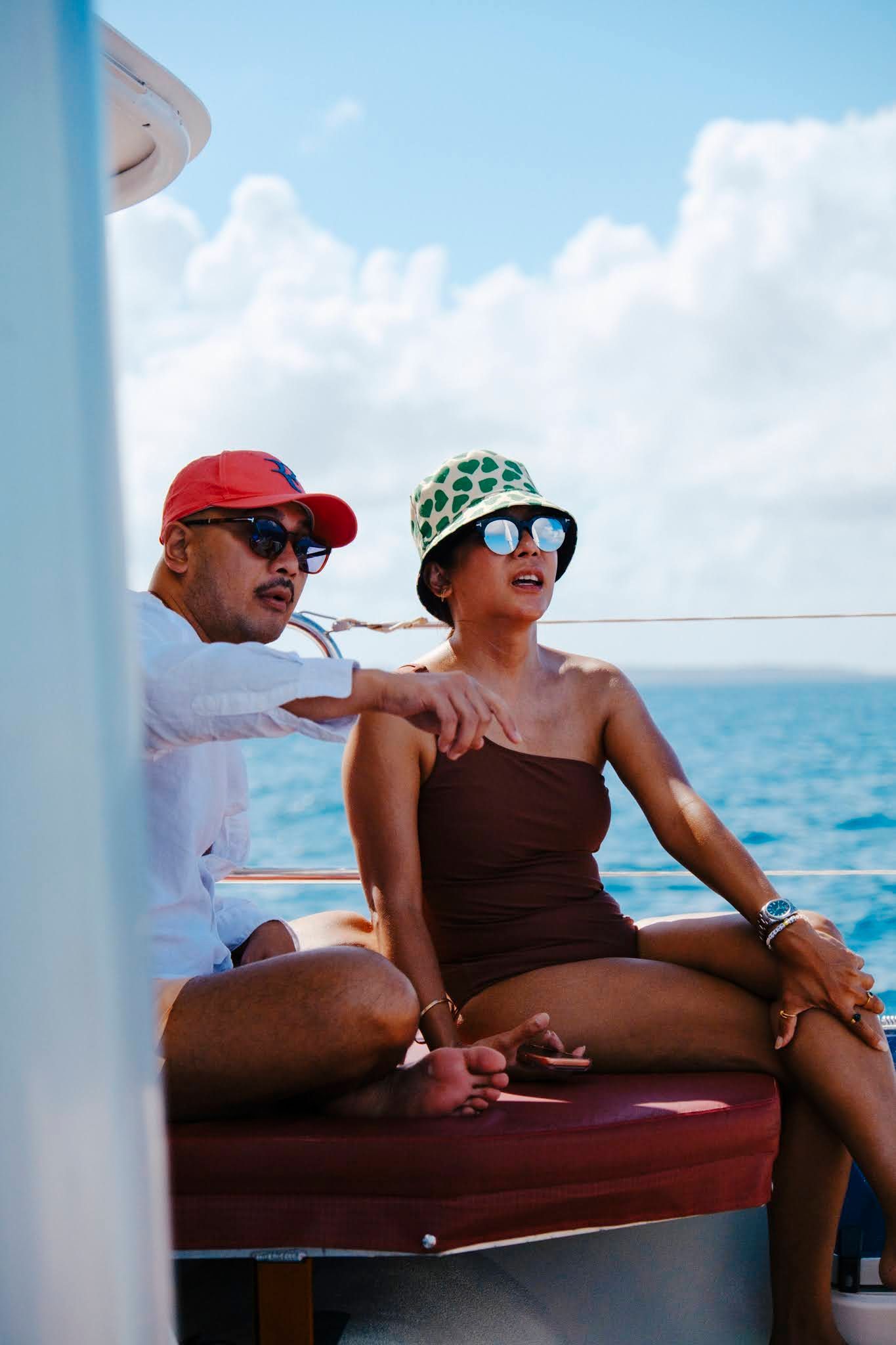 Un couple sur un bateau ; l’homme montre du doigt, la femme lève les yeux. Océan bleu, ciel. Ils portent des chapeaux et des lunettes de soleil.