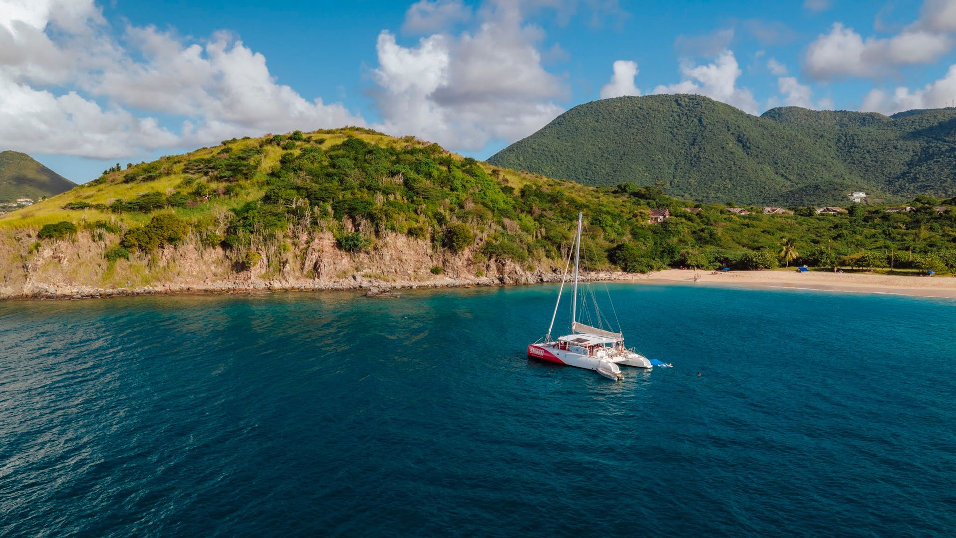 Catamaran sailing on blue water near a beach and green hills.