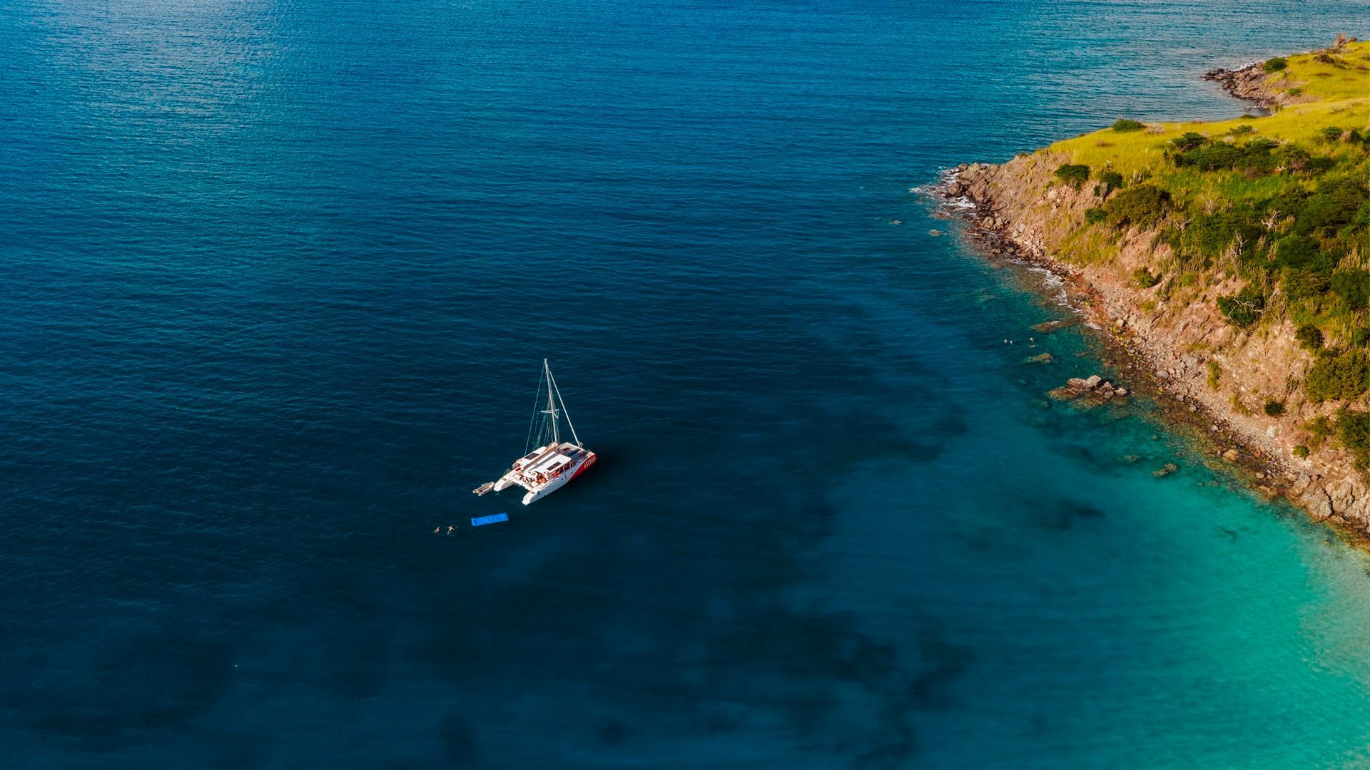 Catamaran sails on turquoise water near a rocky coastline.