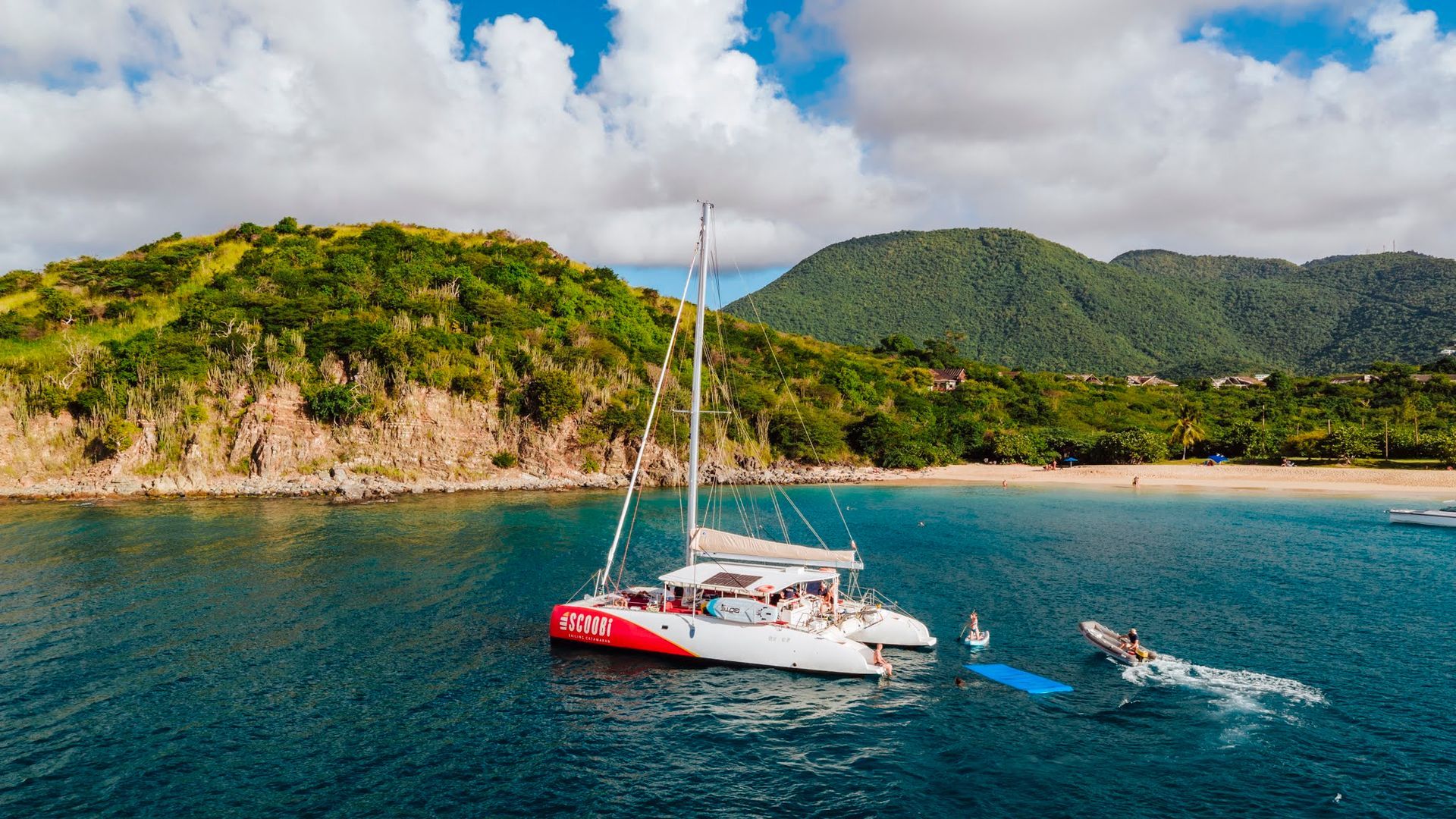Navigation en catamaran près d'une île verdoyante et luxuriante, dotée d'une plage de sable fin, sous un ciel nuageux.
