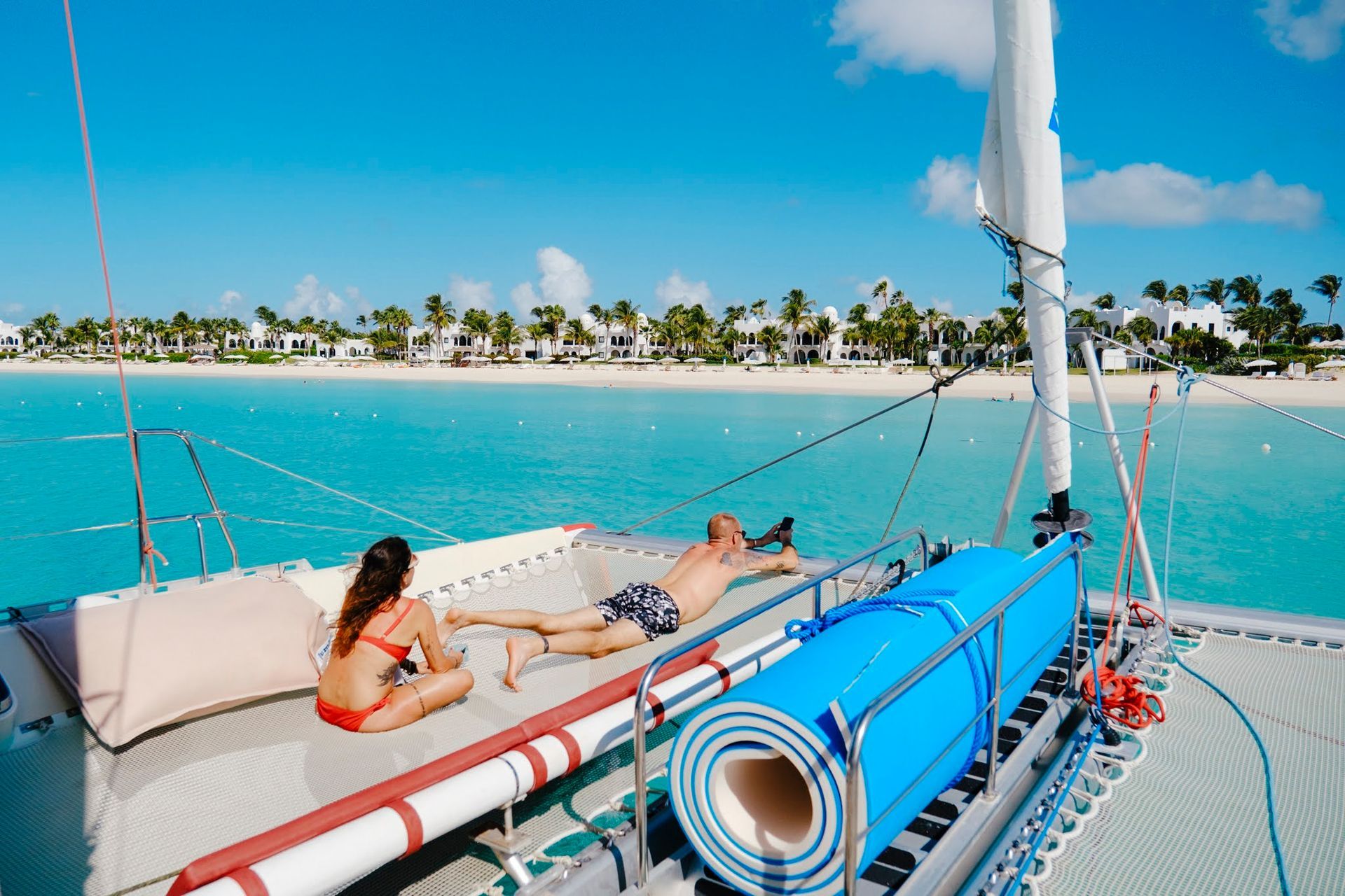 Des personnes prennent un bain de soleil sur un catamaran, eau turquoise, plage de sable blanc et bâtiments en arrière-plan.