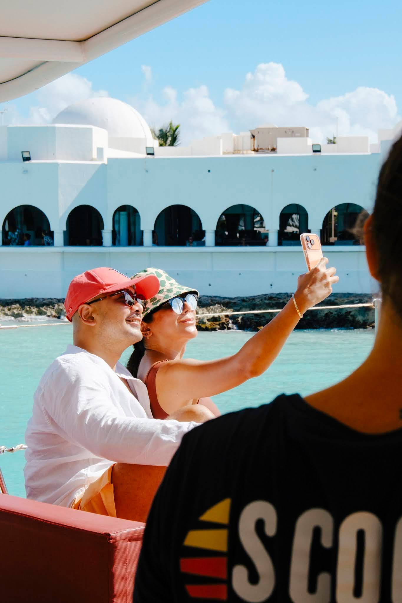 Un couple prend un selfie sur un bateau avec un bâtiment blanc et de l'eau bleue en arrière-plan.