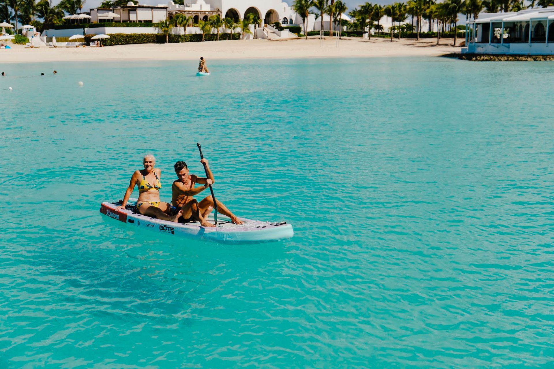 Two people on a paddleboard in turquoise water. White sand beach and buildings in background.