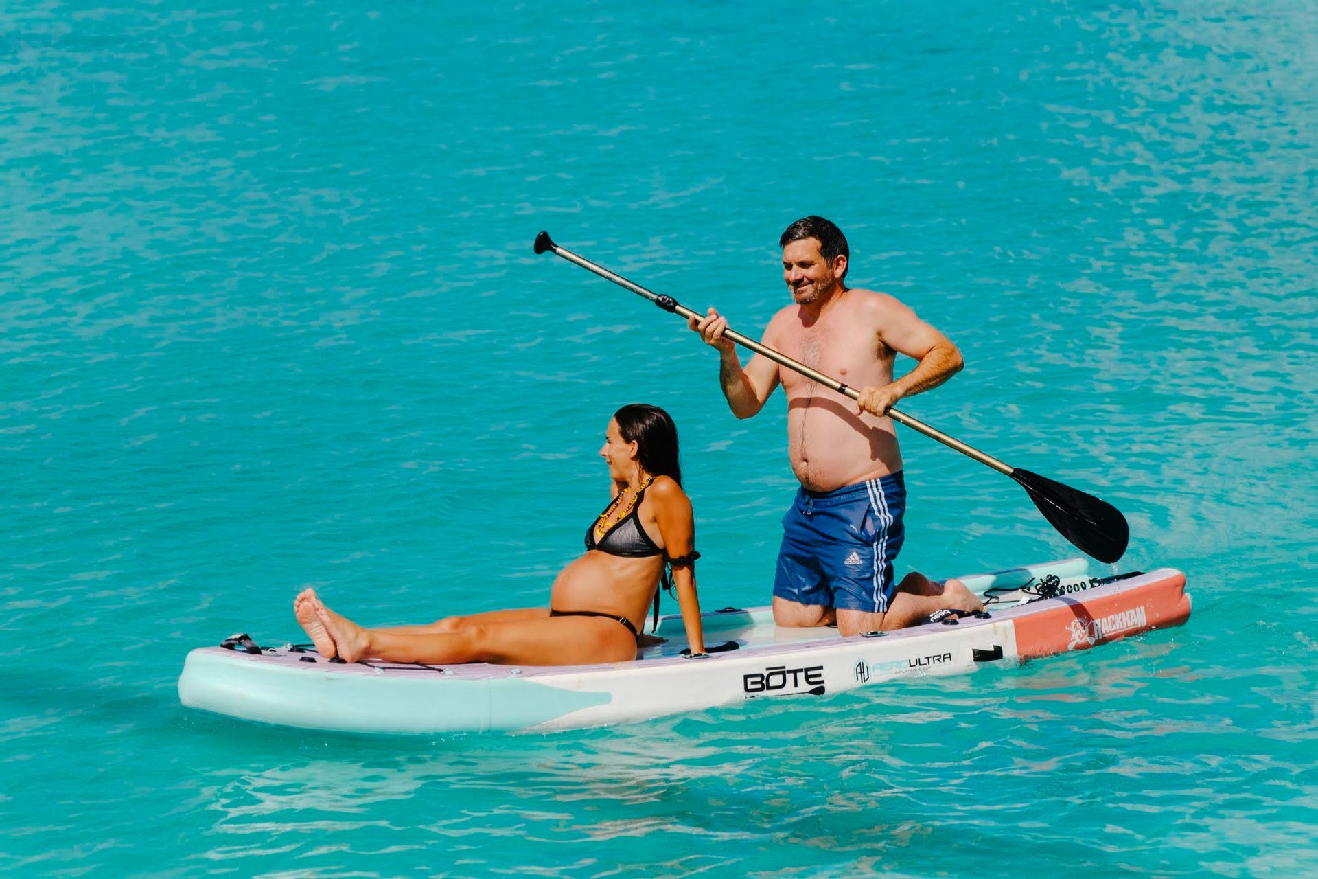 Une femme enceinte et une autre personne sont agenouillées sur une planche à pagaie sur l'eau, l'autre personne tenant une pagaie.