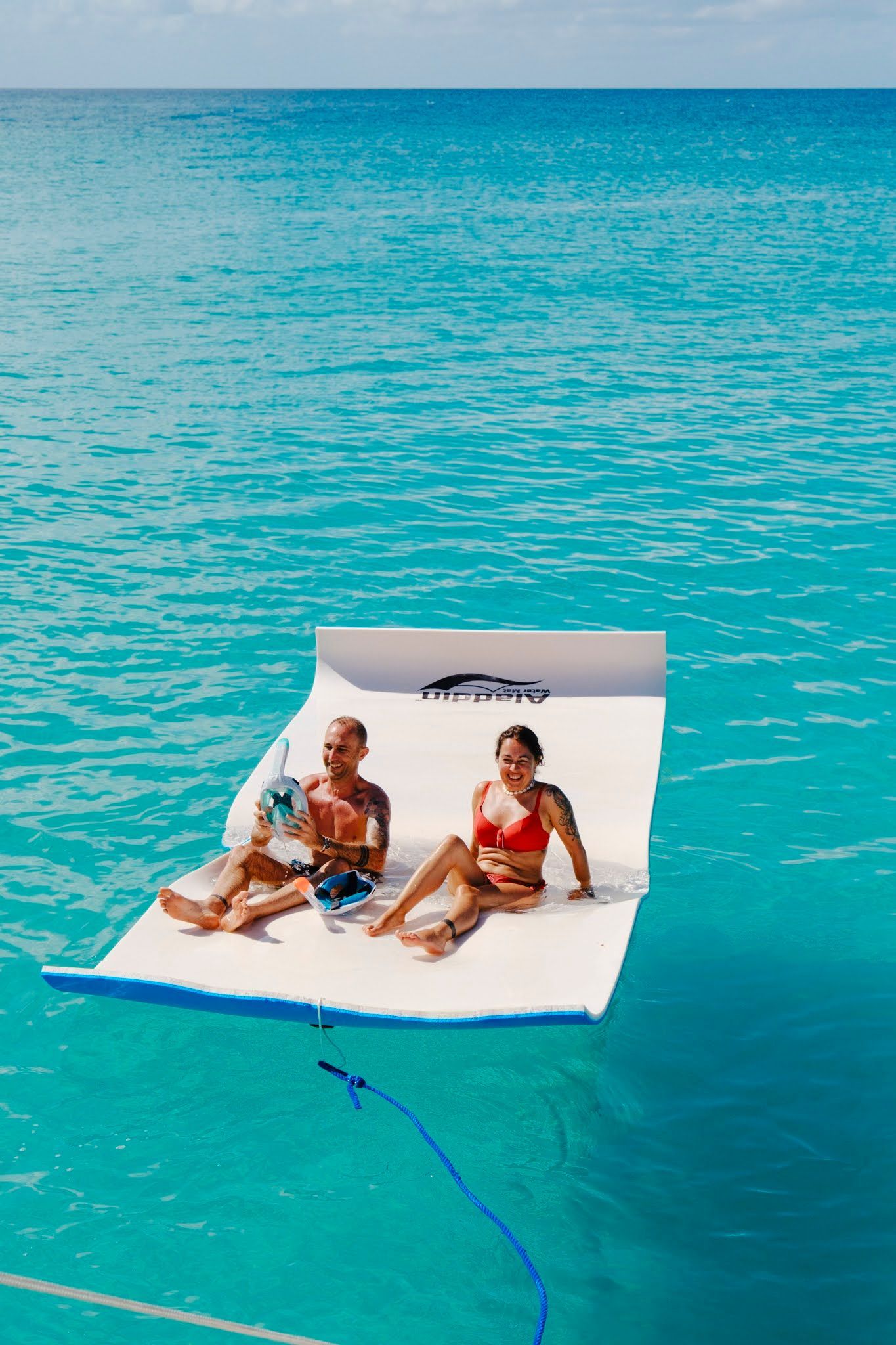 Deux personnes se détendent sur un tapis blanc flottant dans une eau turquoise sous un ciel bleu.