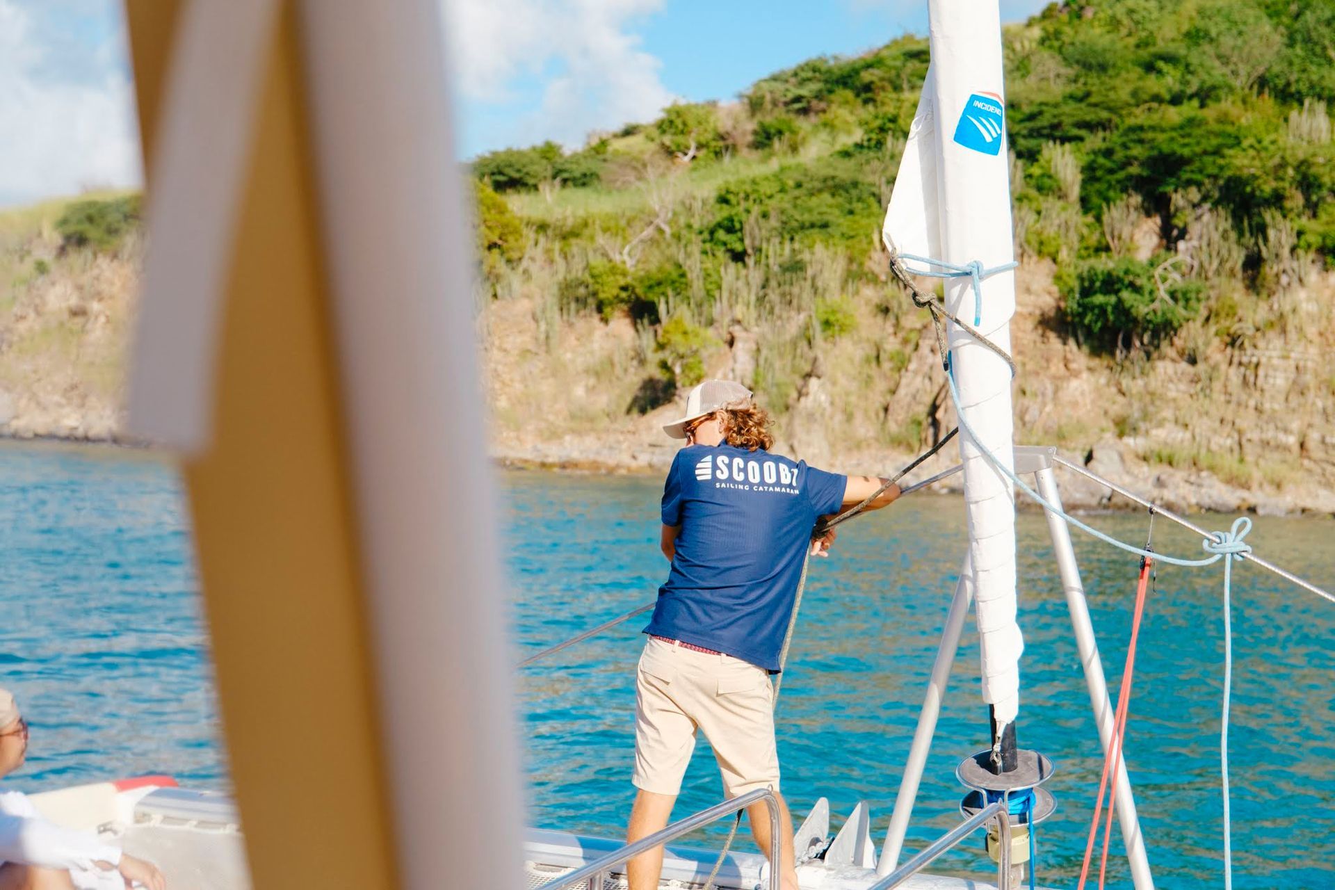 Man on sailboat adjusting sail near a green, hilly shoreline.