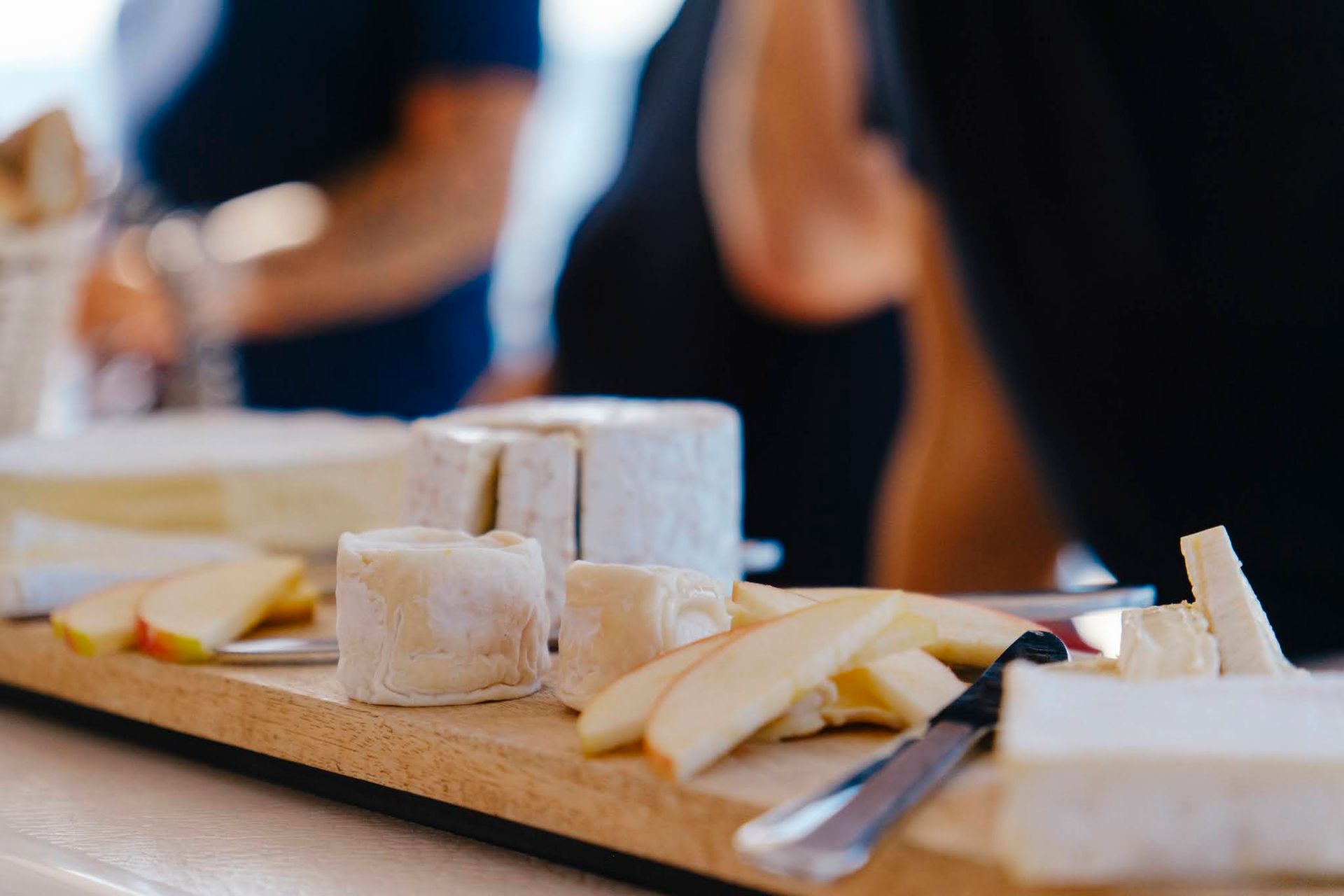 Cheese board with various cheeses and apple slices, with people in the background.