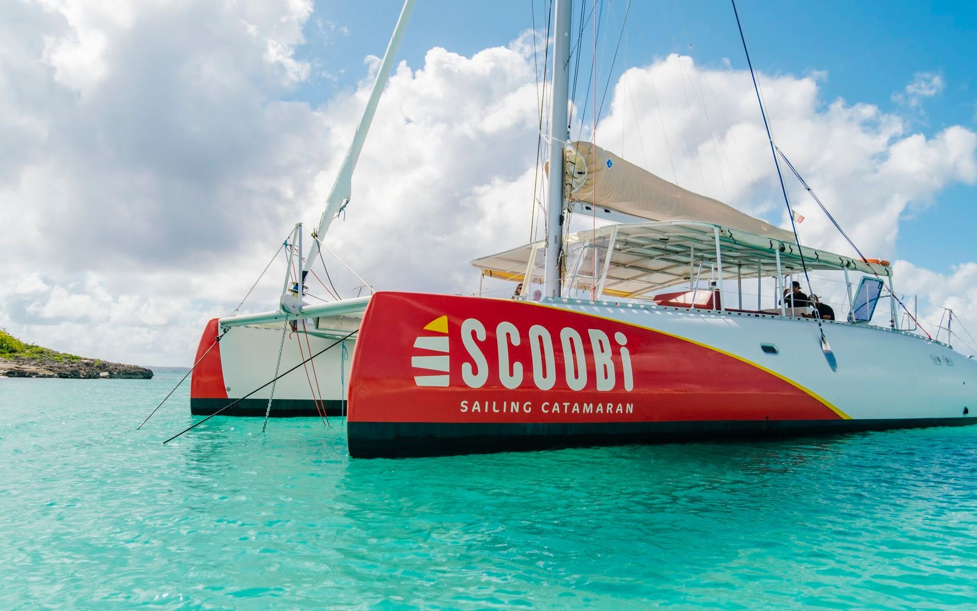 Catamaran « Scoobi » dans une eau turquoise, coque rouge, voiles blanches, ciel ensoleillé.