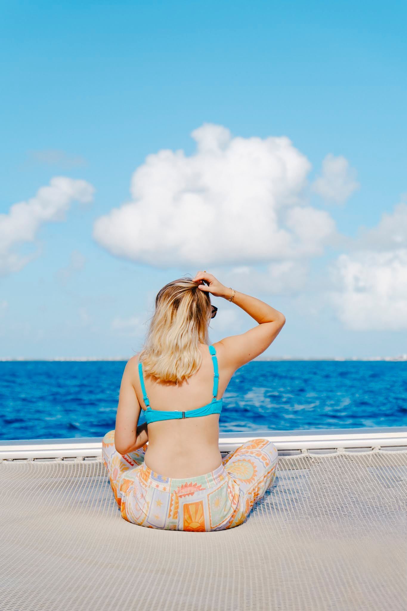 Une femme en haut de bikini turquoise et pantalon à motifs est assise dans un hamac au-dessus de l'océan.