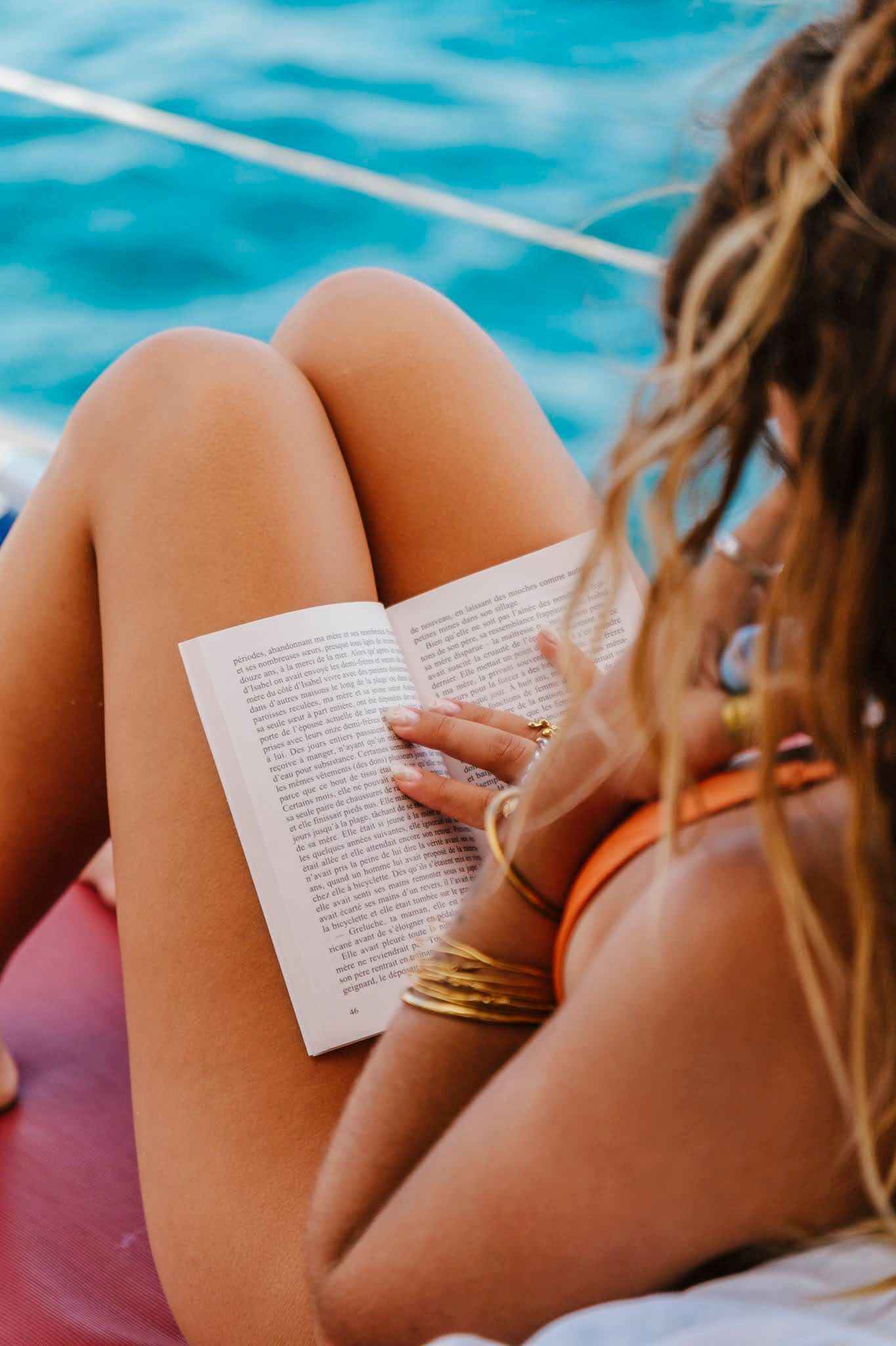 Woman reading a book on a boat, wearing an orange bikini, legs crossed, against a blue sea.