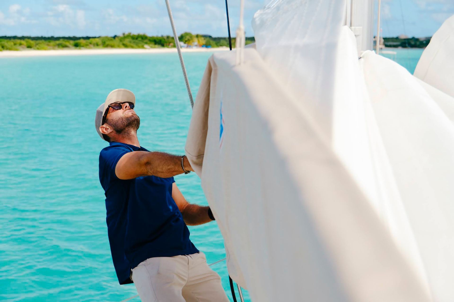 Un homme sur un voilier tire sur une voile, face à une eau turquoise et limpide. Journée ensoleillée.