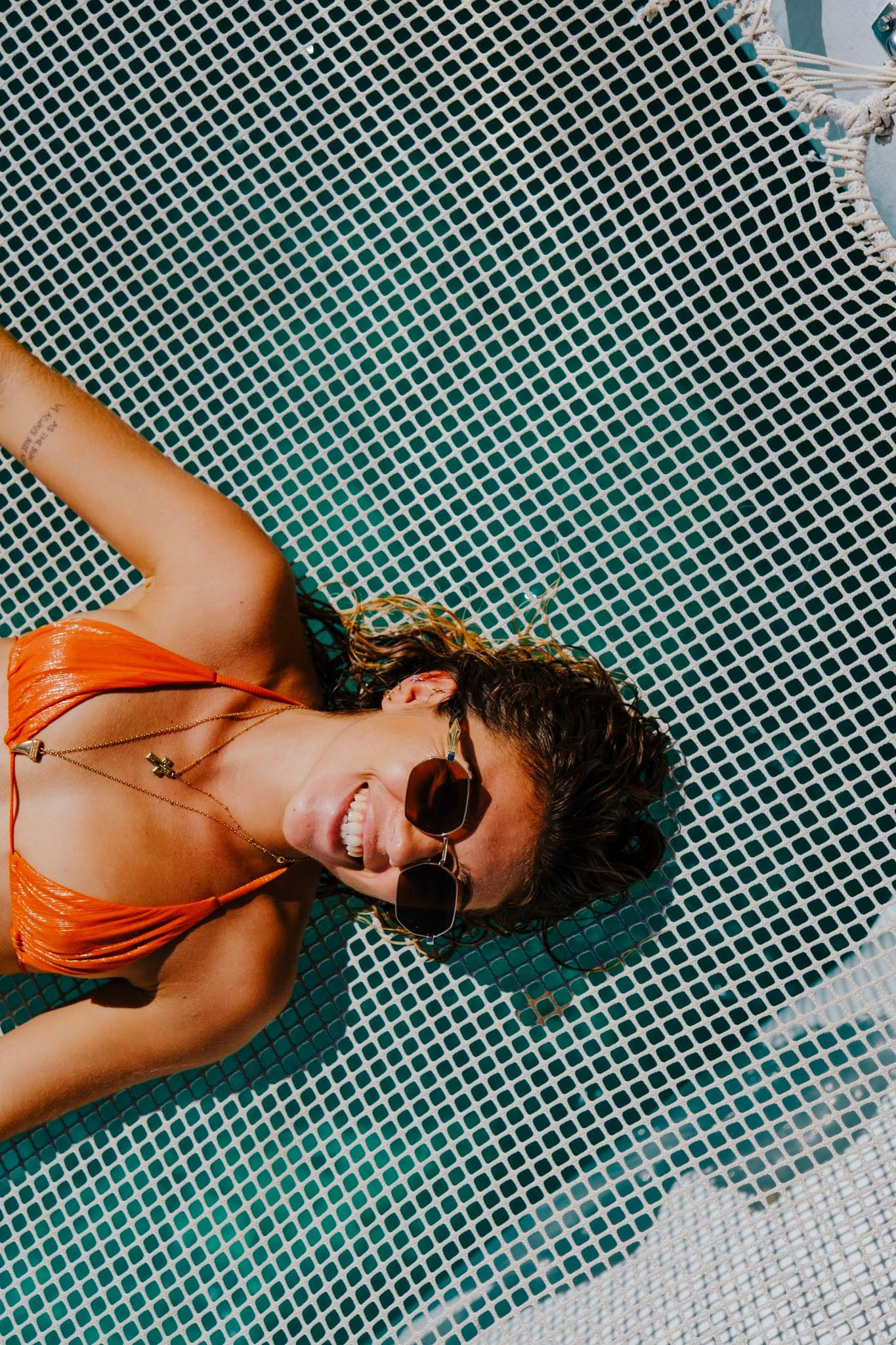 Woman in orange bikini, sunglasses, smiles, relaxed in a hammock over turquoise water.