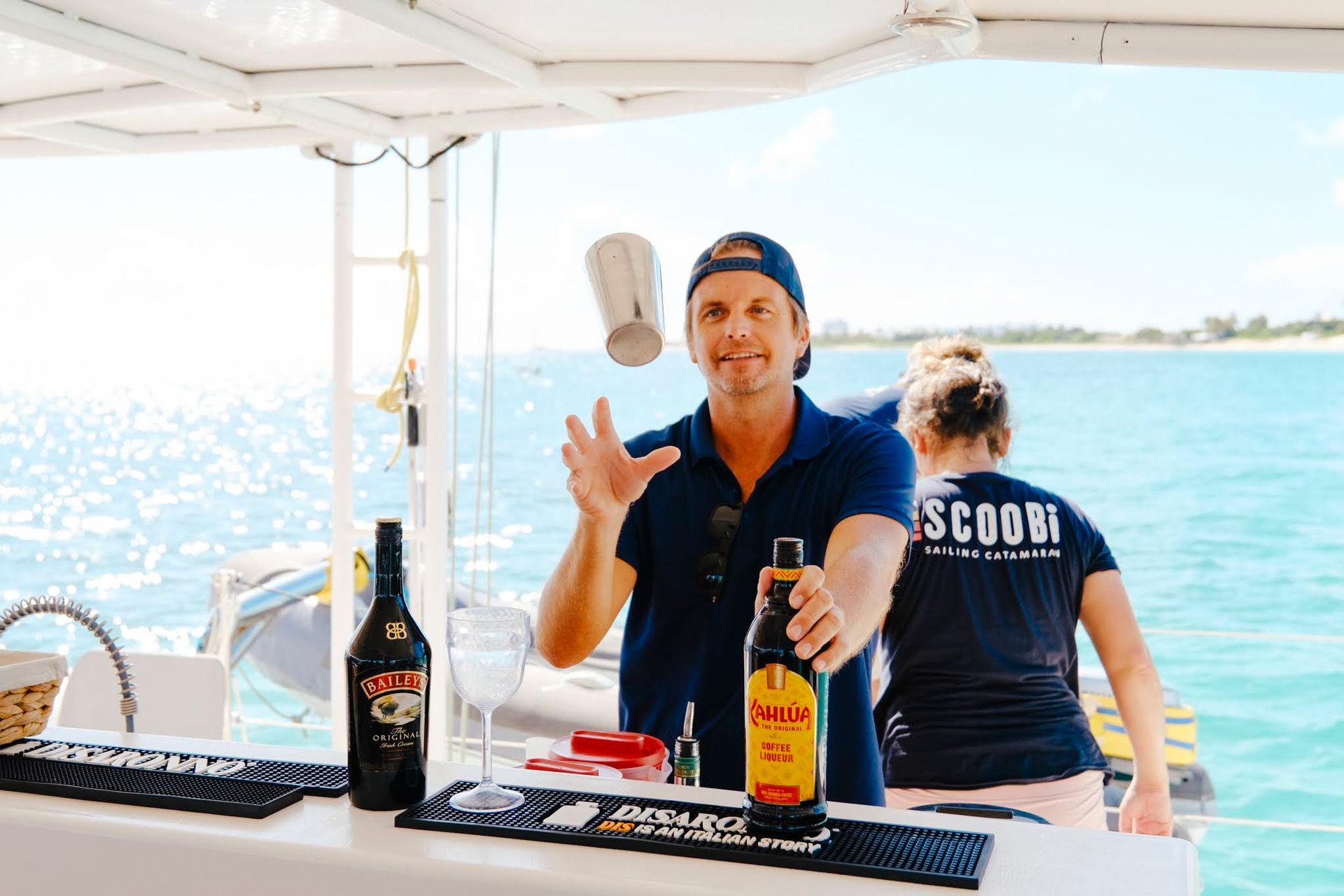 Man juggling a cocktail shaker behind a bar on a boat; Baileys and rum bottles visible. Ocean in background.