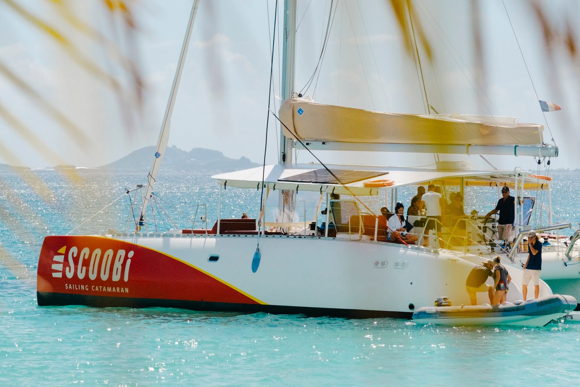 Le catamaran « Scoobi » en mer avec des personnes à bord. Bateau rouge et blanc à voiles beiges par une journée ensoleillée.
