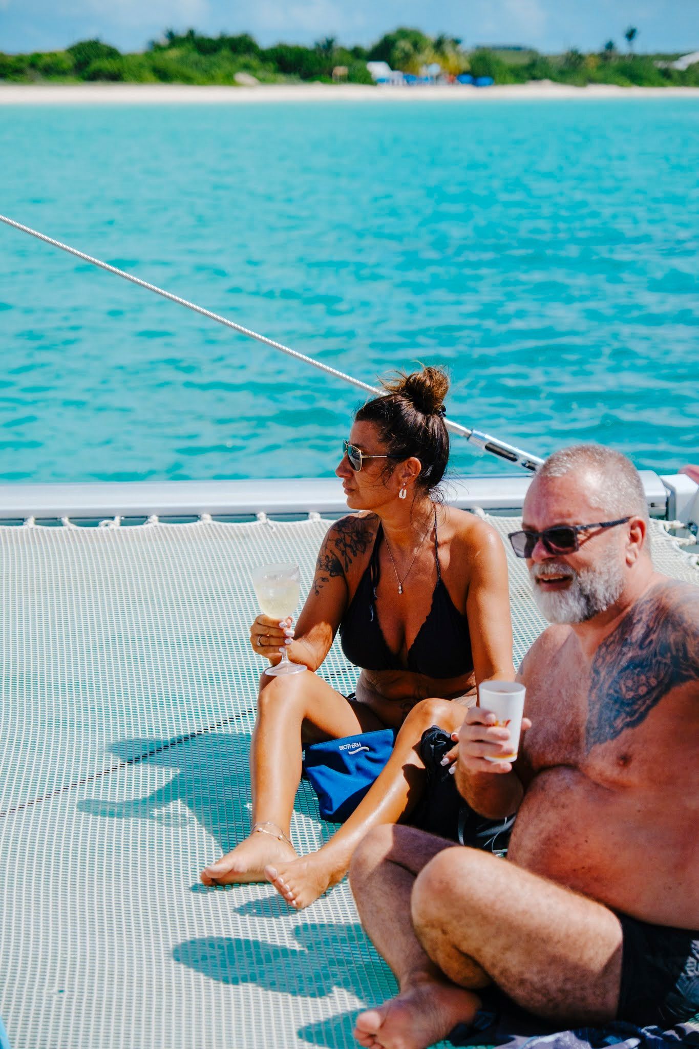 Un couple se détend sur un catamaran, en sirotant des boissons, avec l'océan et la plage en arrière-plan.