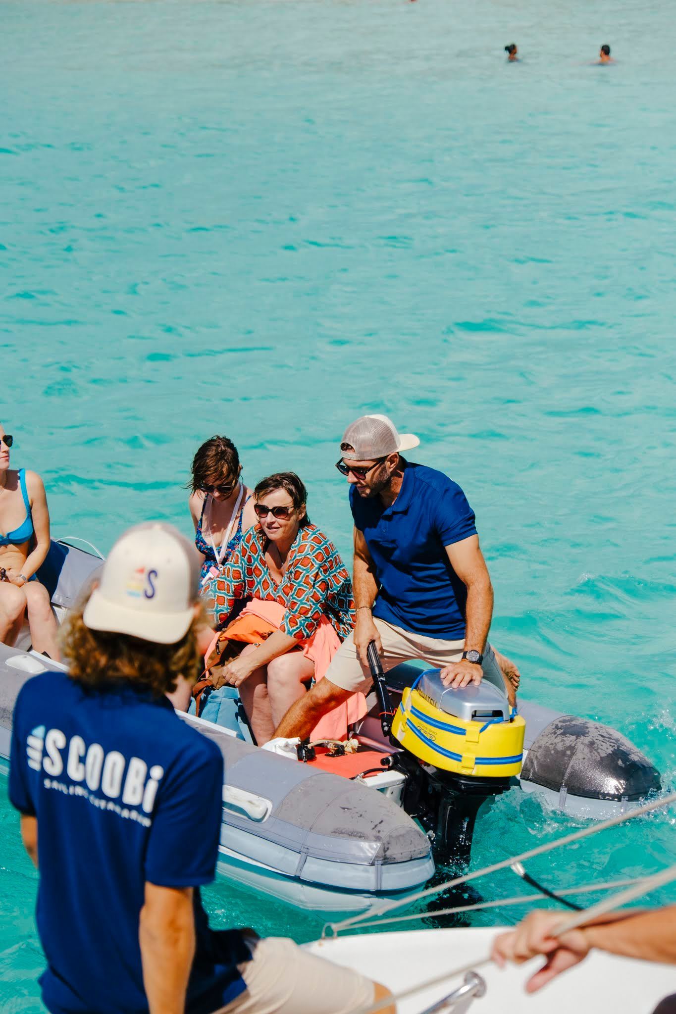 Des personnes se trouvent à bord d'un petit bateau pneumatique sur une eau turquoise. L'une d'elles manœuvre le moteur hors-bord, les autres sont assises.