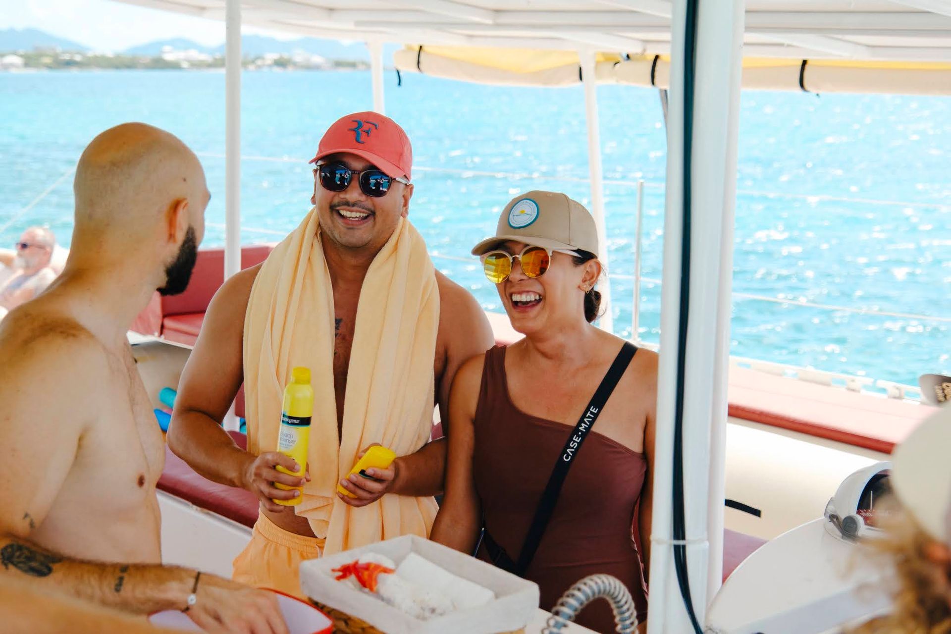 People on a boat smiling. One holds a drink, the woman laughs, set against the ocean.