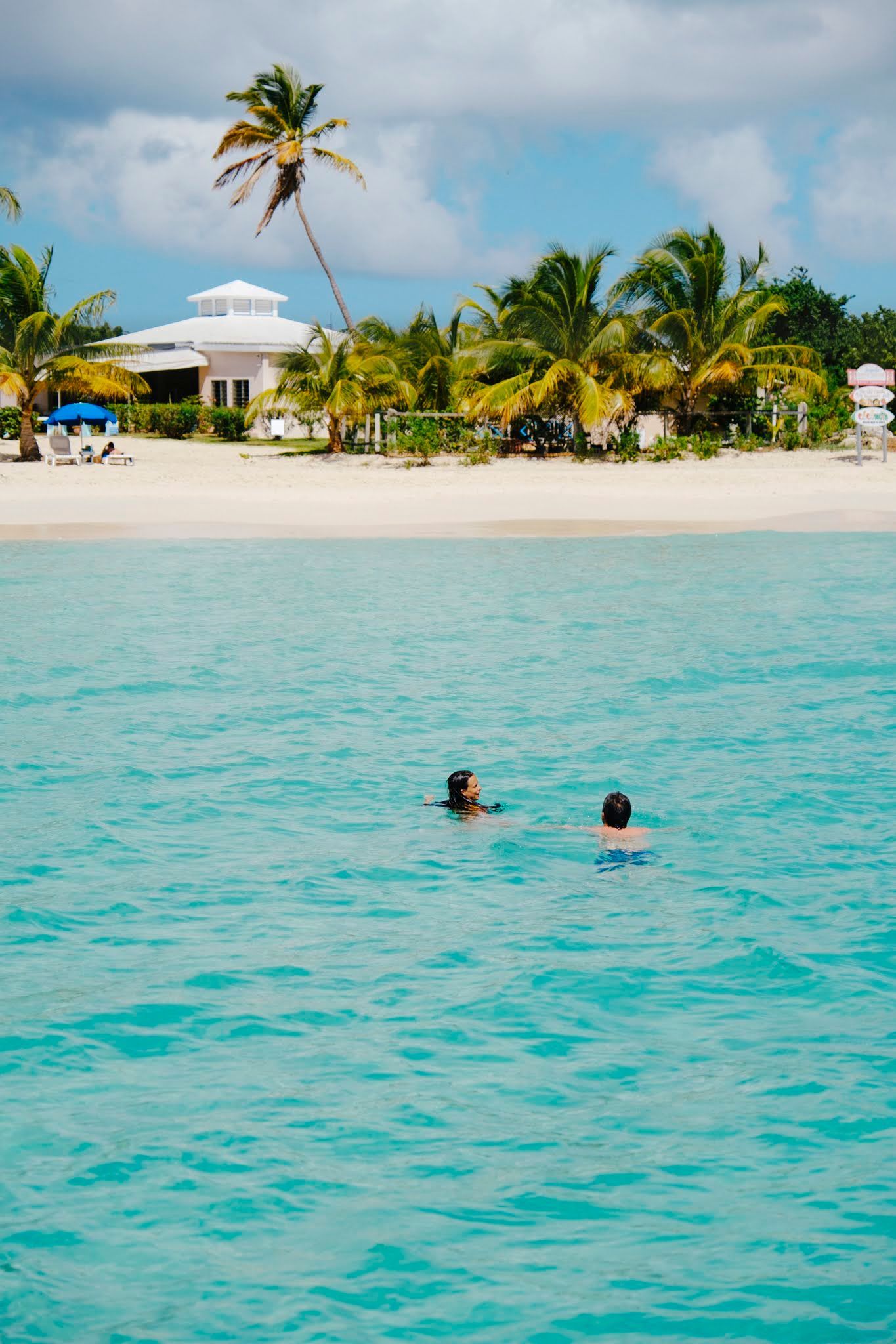 Two people swimming in turquoise water near a white sandy beach with palm trees and buildings.