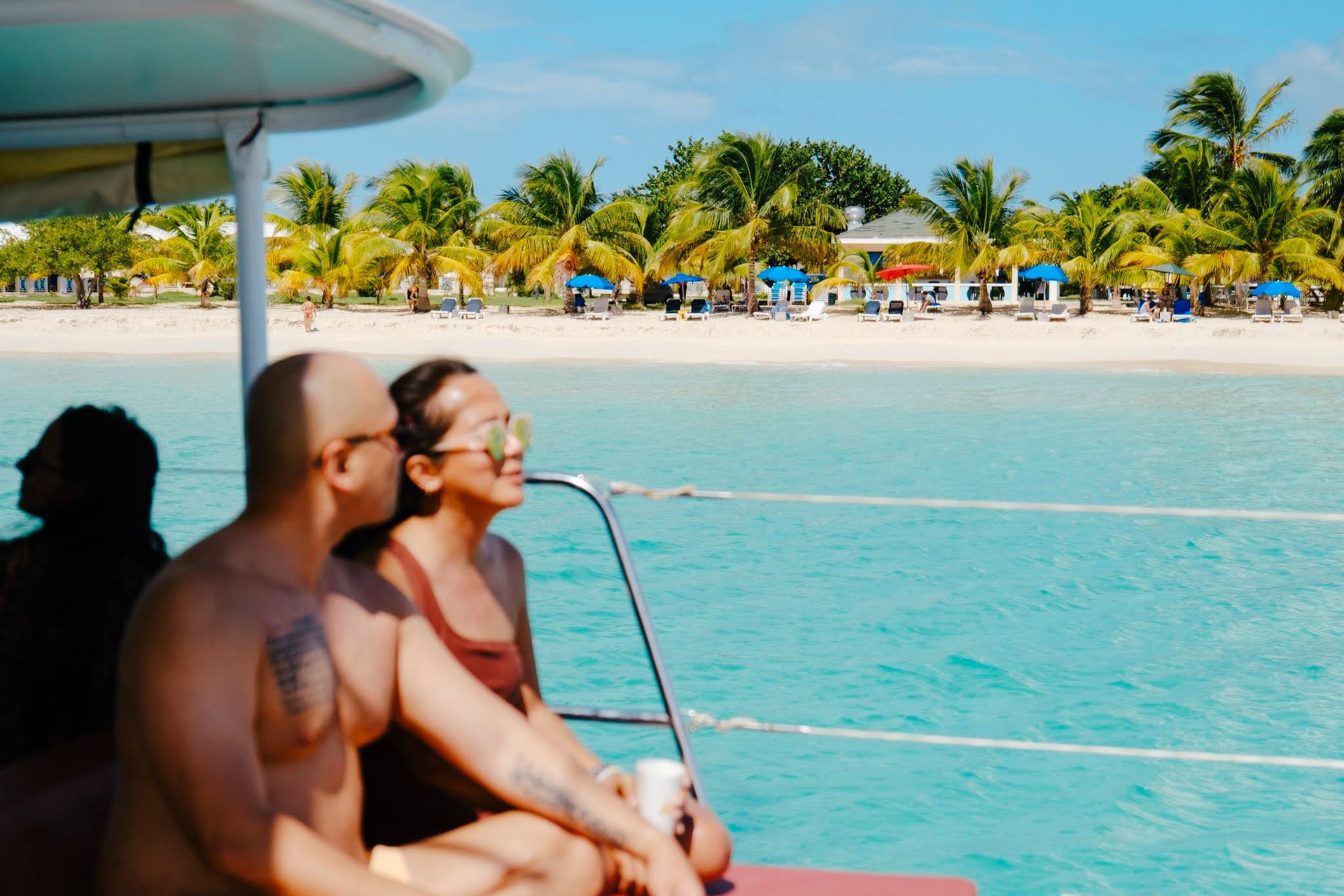 Two people on a boat near a tropical beach. Turquoise water, sunny day, trees, white sand.