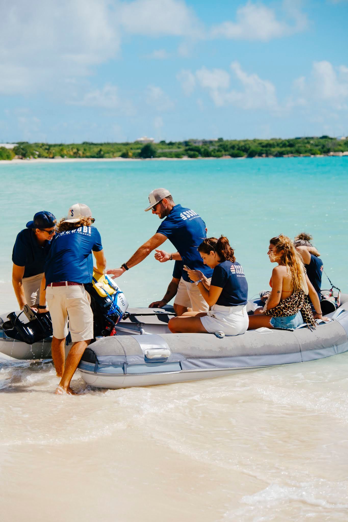 People disembarking a boat onto a sandy beach.  Crew members assist passengers near turquoise water under a blue sky.