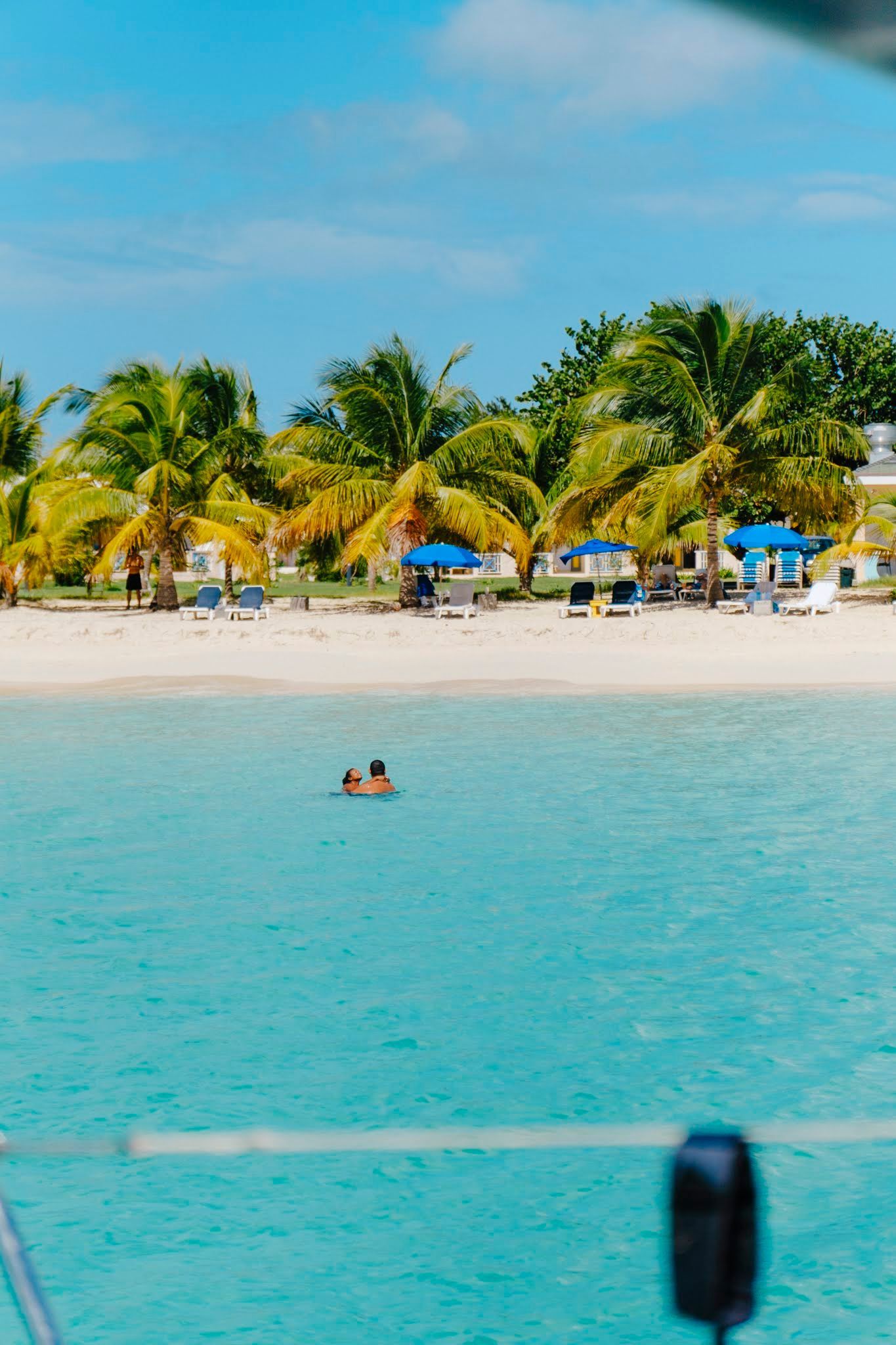 Bright turquoise water with a sandy beach lined with palm trees and blue umbrellas. Two people swim in the water.
