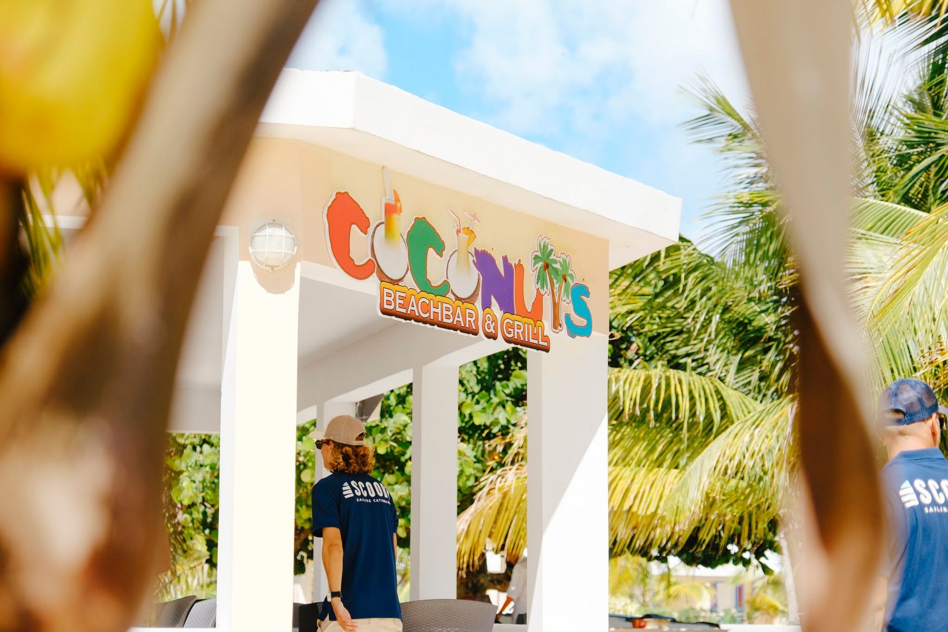 Panneau indiquant le Coconuts Beach Bar, avec une personne derrière le comptoir et des palmiers en arrière-plan.