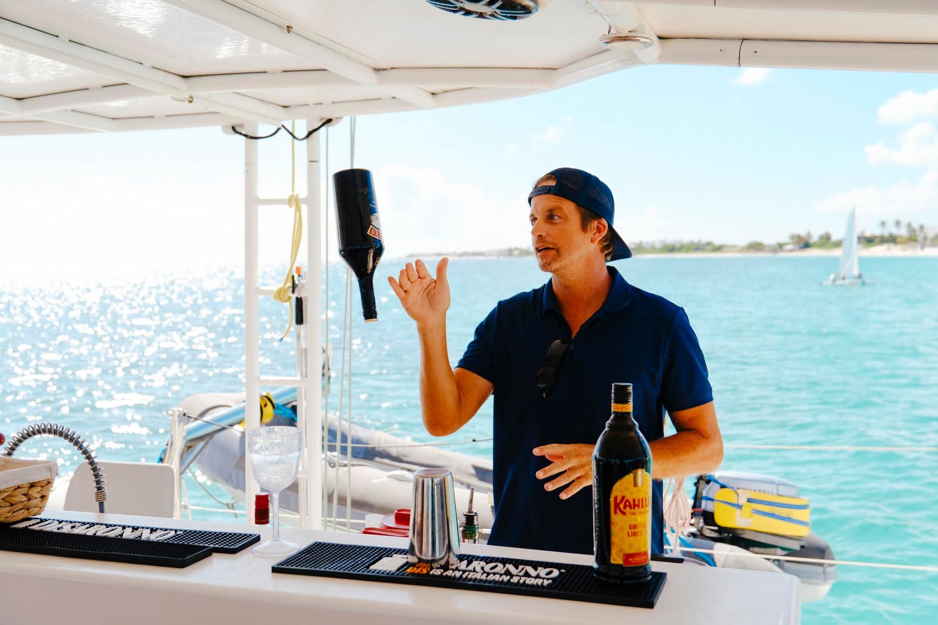 Man on a boat, pouring a bottle of wine. Blue water and sky.